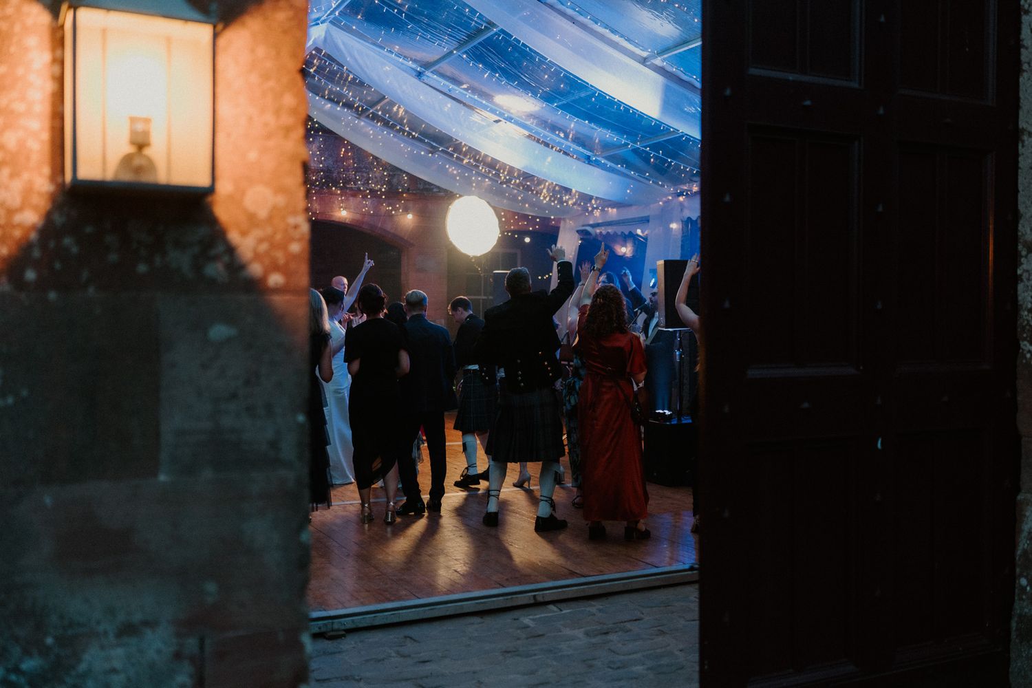 Wedding guests in formal attire dance with raised arms in a candlelit reception venue, seen through a rustic stone doorway at night.