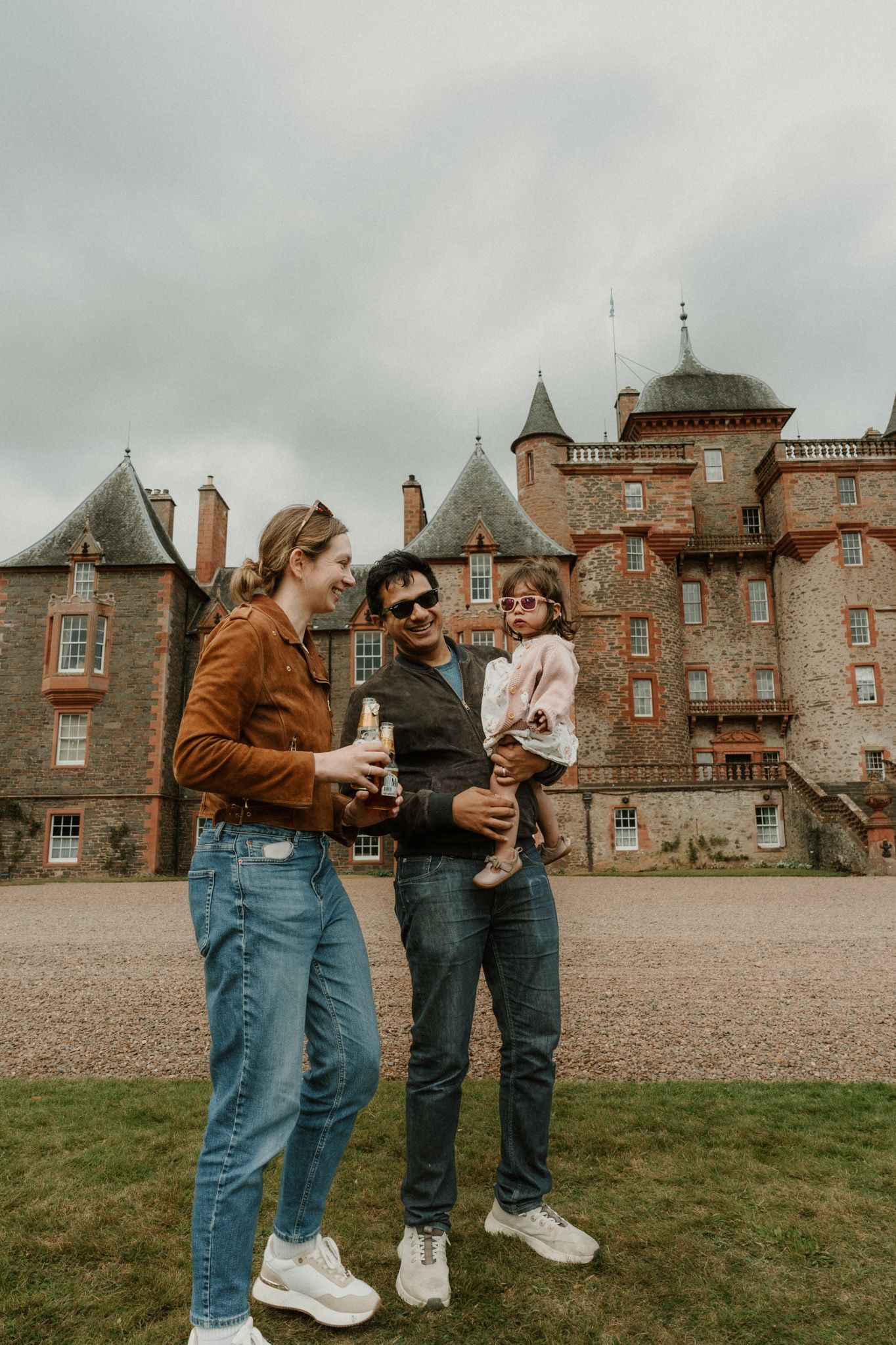 A smiling woman and a man holding a toddler stand on the lawn in front of a historic stone castle with turrets under a cloudy sky.