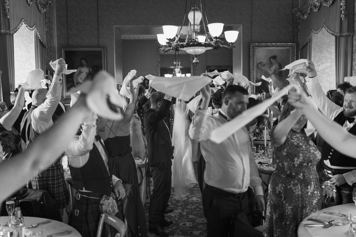 Wedding guests in a grand ballroom wave white napkins overhead in black and white as a chandelier hangs above.