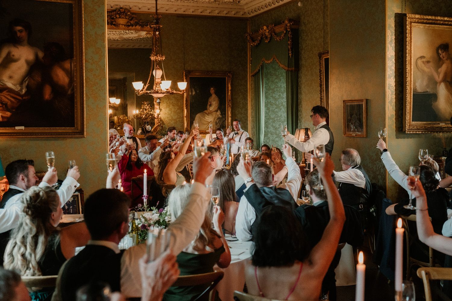 Wedding guests raise glasses and cheer in a grand dining room with chandeliers, ornate gold-framed paintings, and lit candles on the tables.