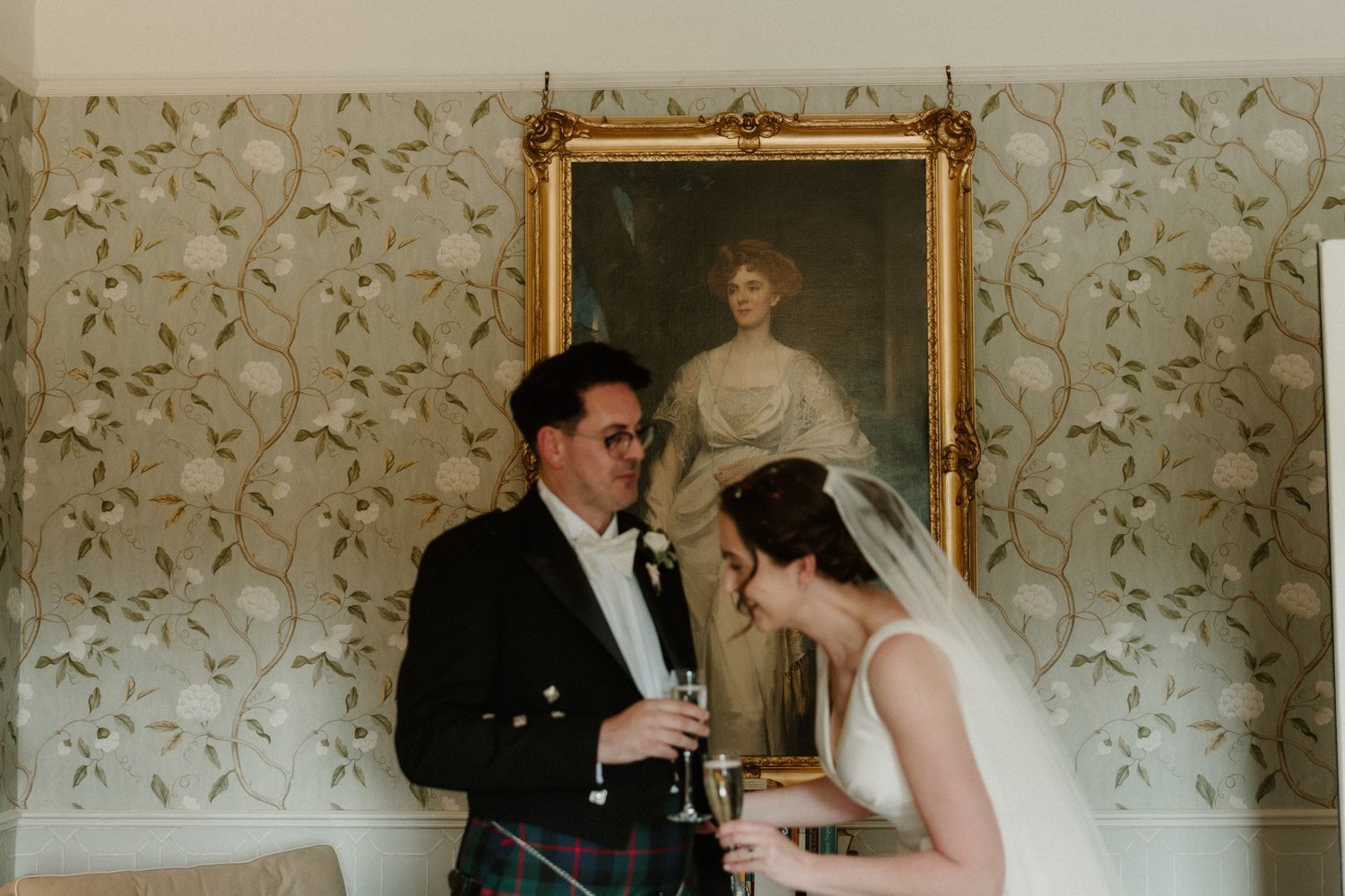 Bride in a veil and groom in a Scottish kilt jacket share a champagne toast beneath a gilded framed portrait on a floral wallpapered wall.