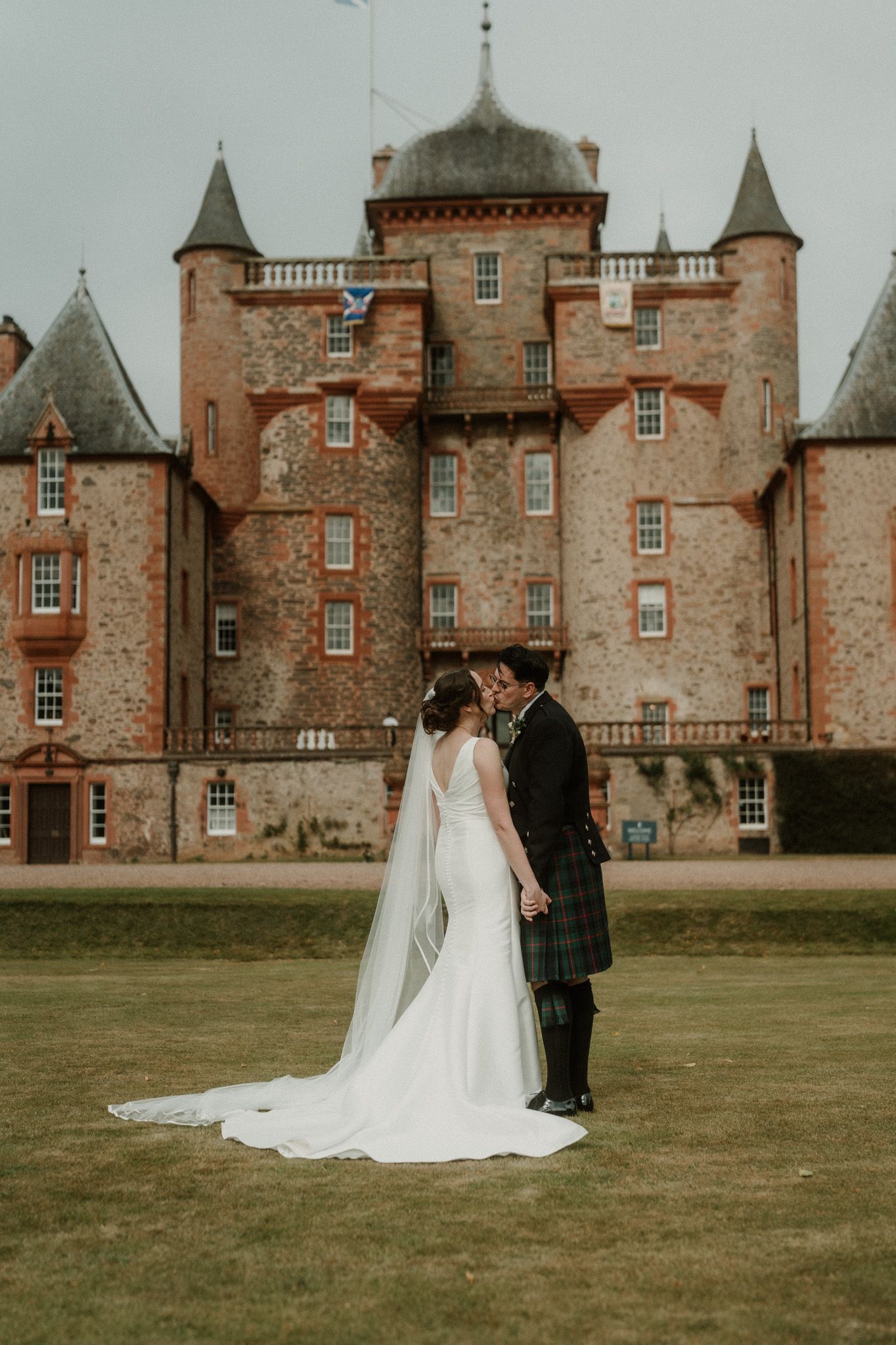 Bride in a white gown and groom in a kilt share a kiss on the lawn in front of a red sandstone Scottish castle.