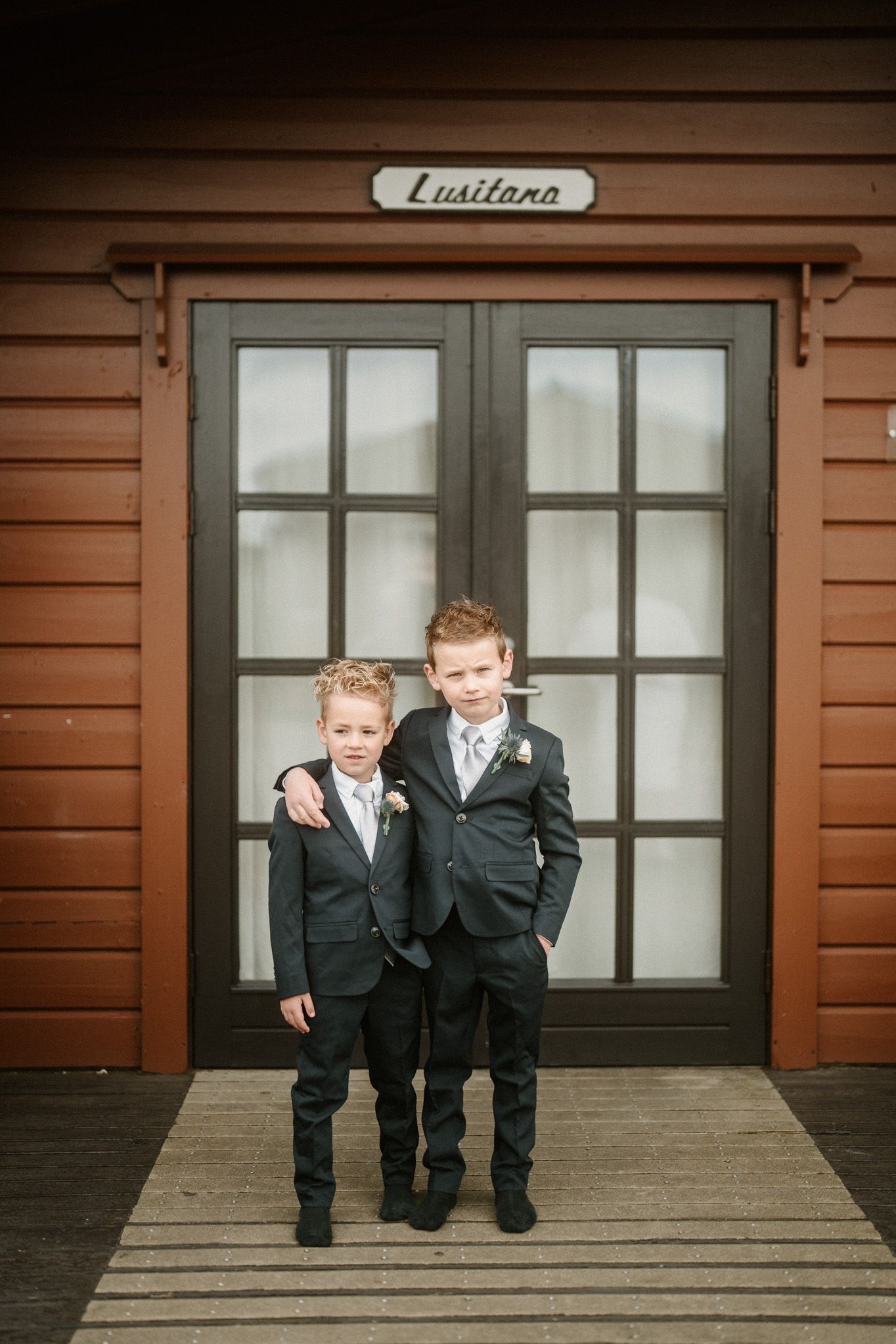 Two young boys in formal suits playing outside a wooden building with windows.