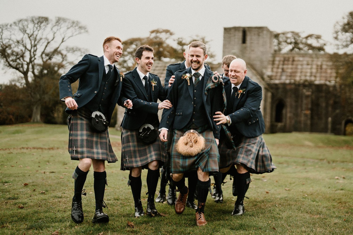Wedding party in traditional Scottish attire walks across green field near historic ruins.