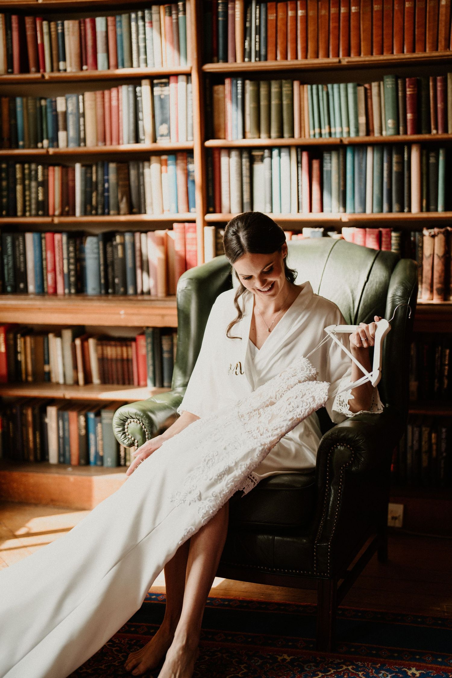 A white wedding dress is displayed in a cozy library setting with wall-to-wall bookshelves.