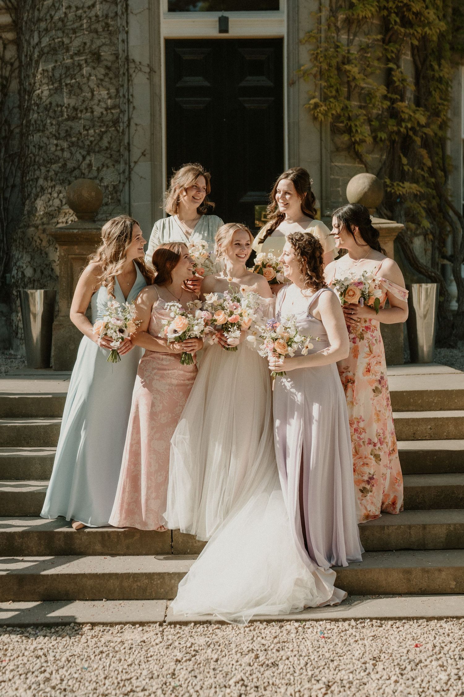 Bridal party in pastel dresses posing with bouquets on stone steps.