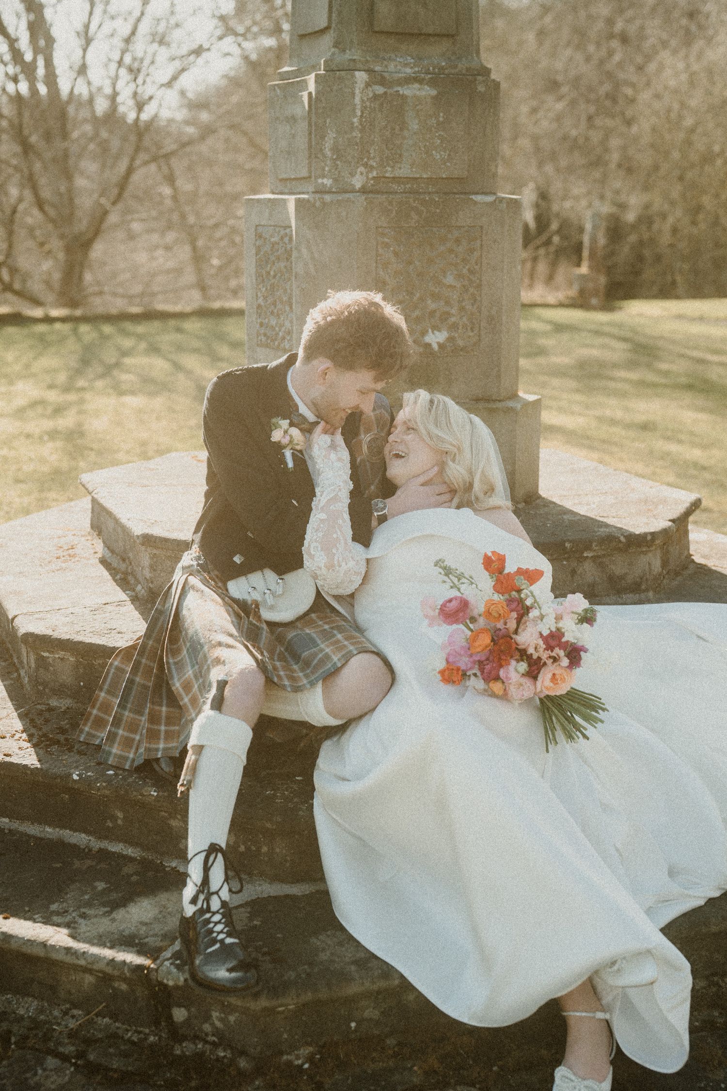 Romantic wedding couple shares a tender moment on stone steps with the groom in traditional Scottish dress.