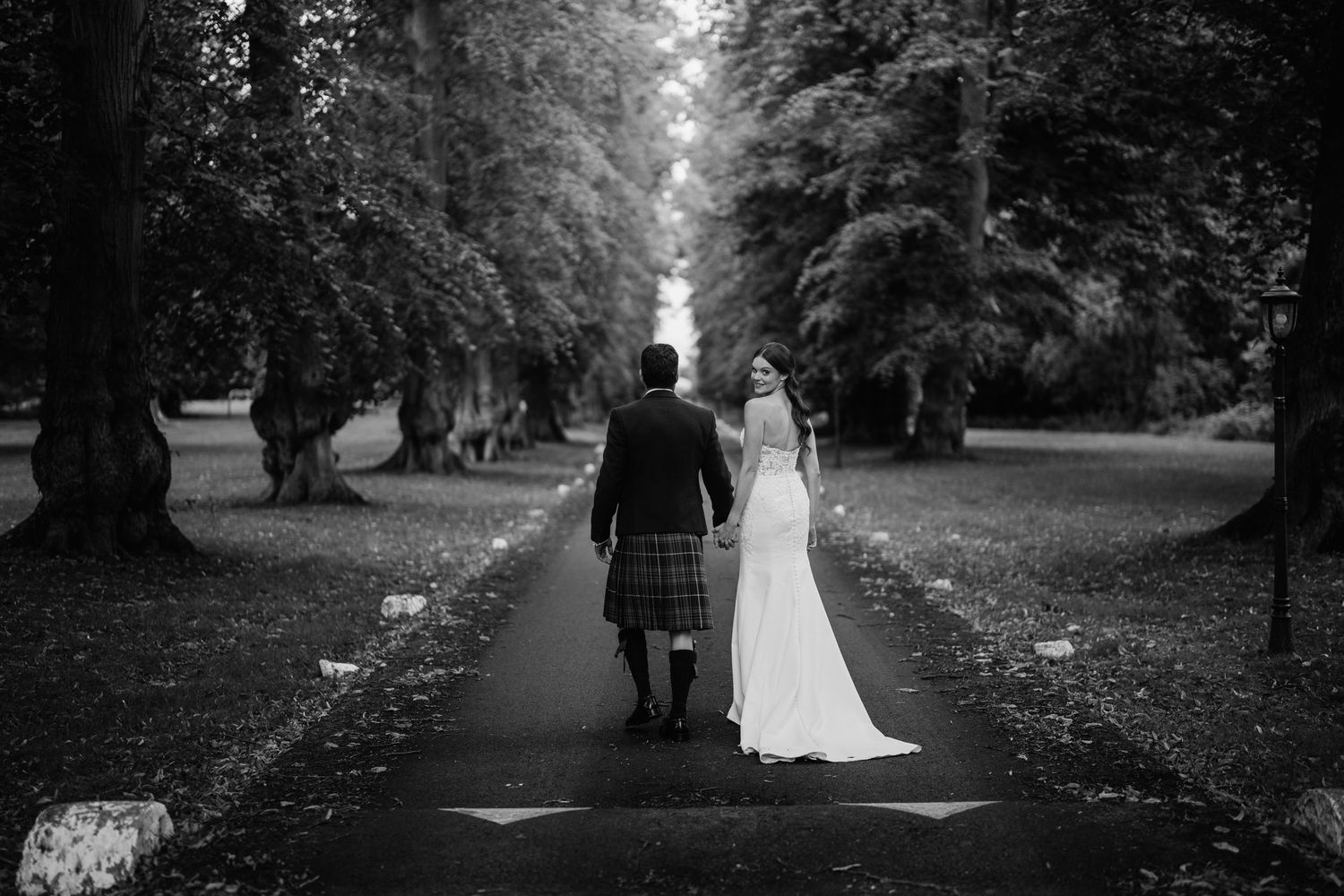 Black and white photo of a tree-lined path in a scenic park setting.
