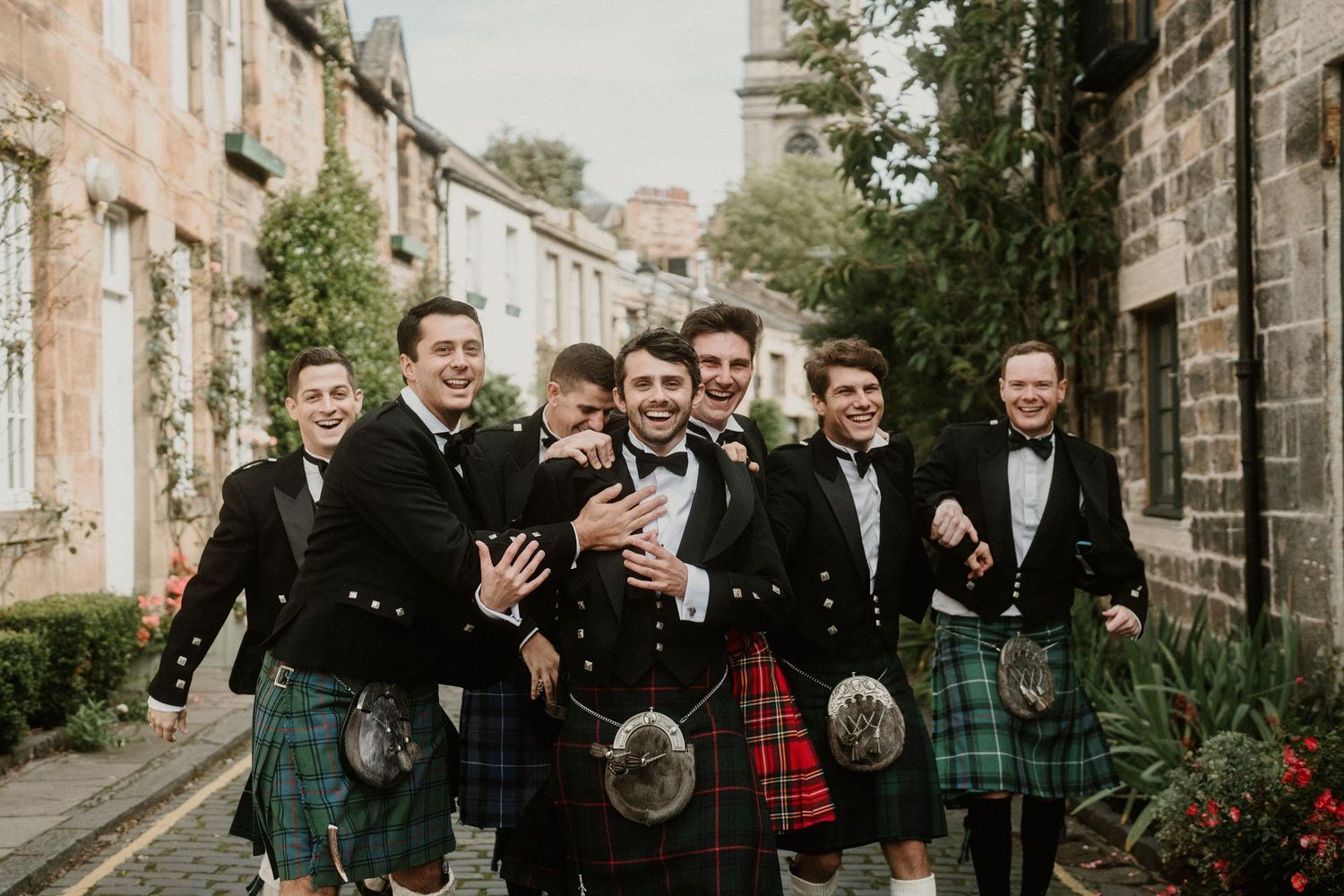 Groomsmen in kilts laugh together while walking down charming stone-lined street.