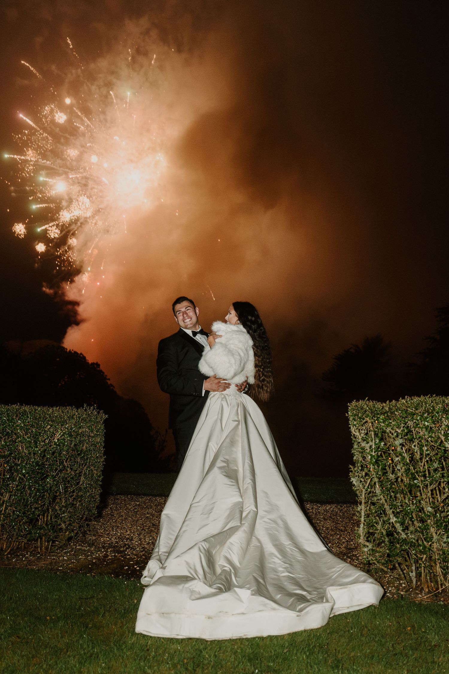 Dramatic fireworks illuminate the night sky behind couple in wedding attire.