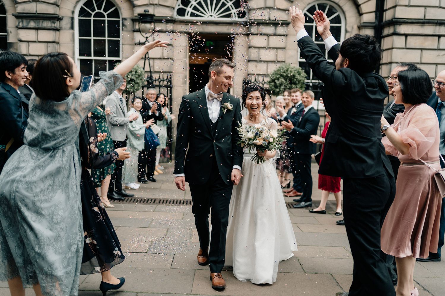 Wedding party and guests gather outside a classical stone building for photographs.