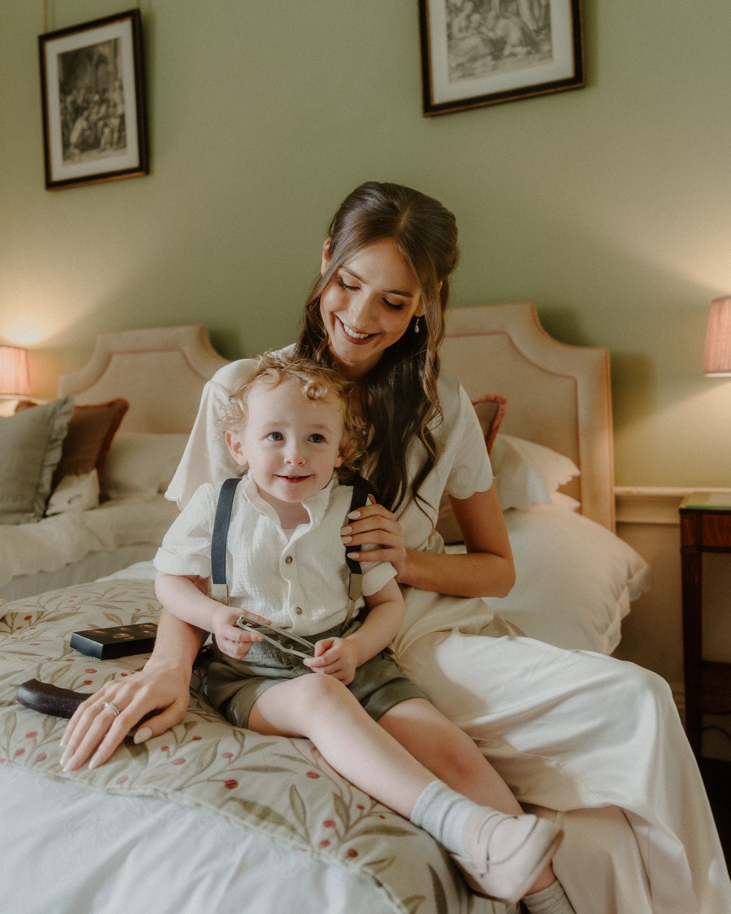 A smiling bride in white dress sits on bed with curly-haired toddler boy in suspenders in a green bedroom.
