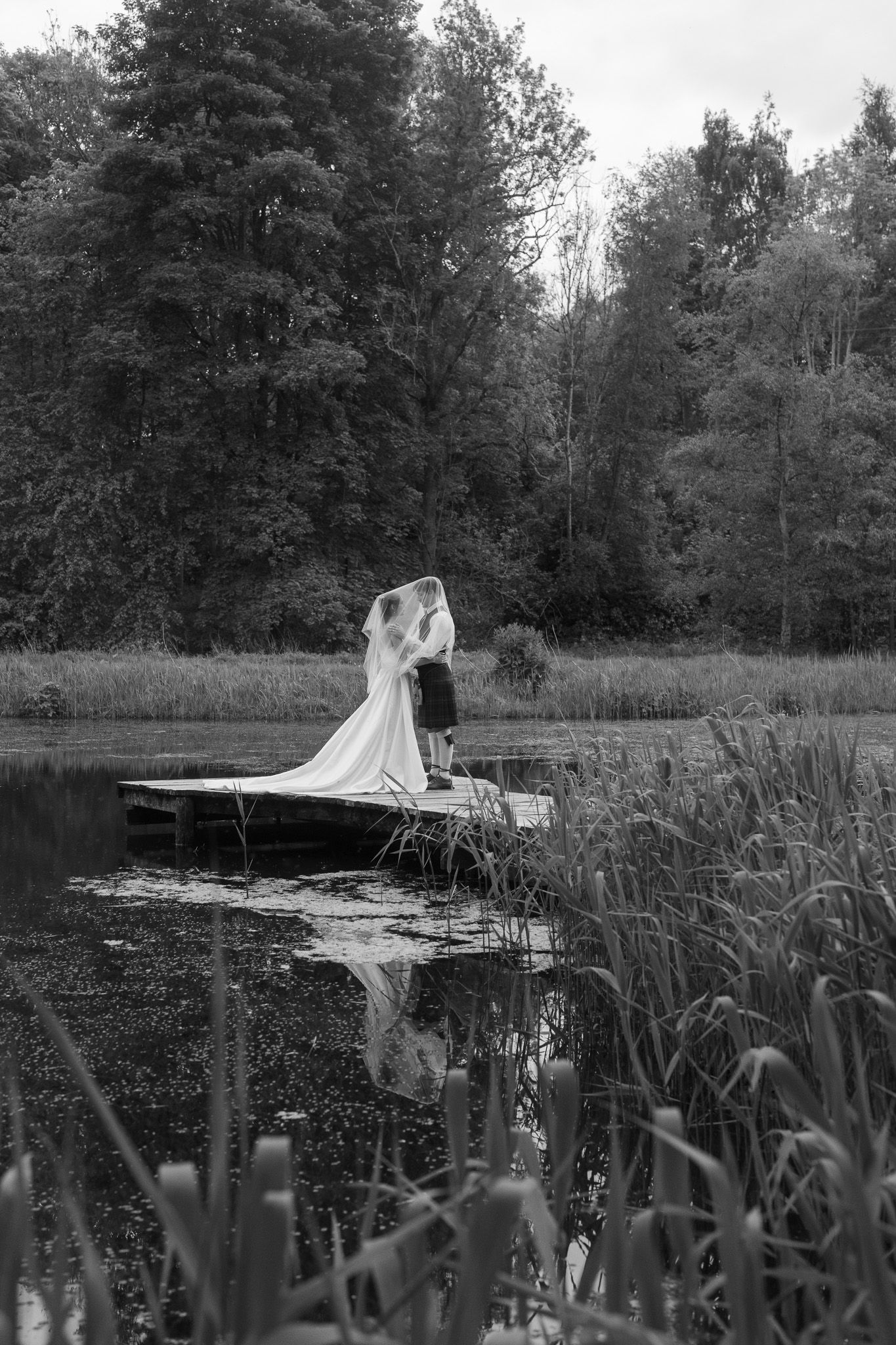 Black and white photo of bride and groom kissing on a wooden dock surrounded by a tranquil lake and lush trees.