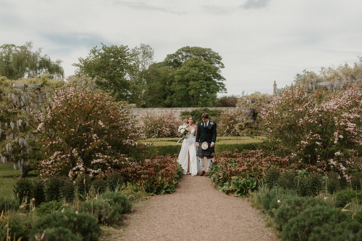 Bride and groom stand together on a garden path surrounded by blooming flowers at an elegant outdoor wedding venue.
