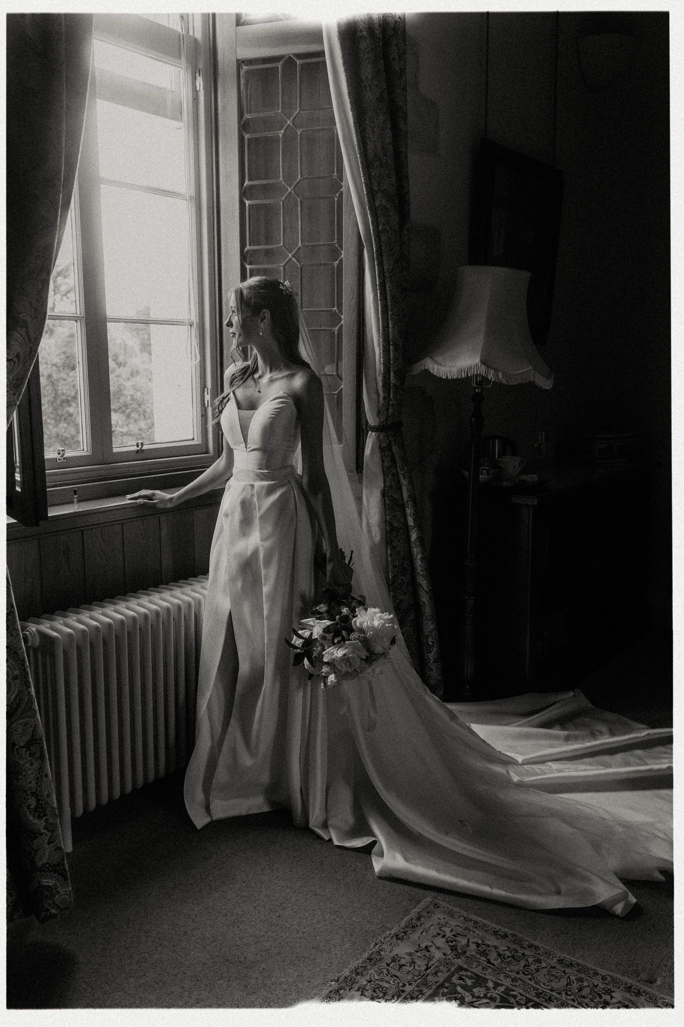 Black and white photo of bride in sleek wedding dress with train standing by leaded glass window.