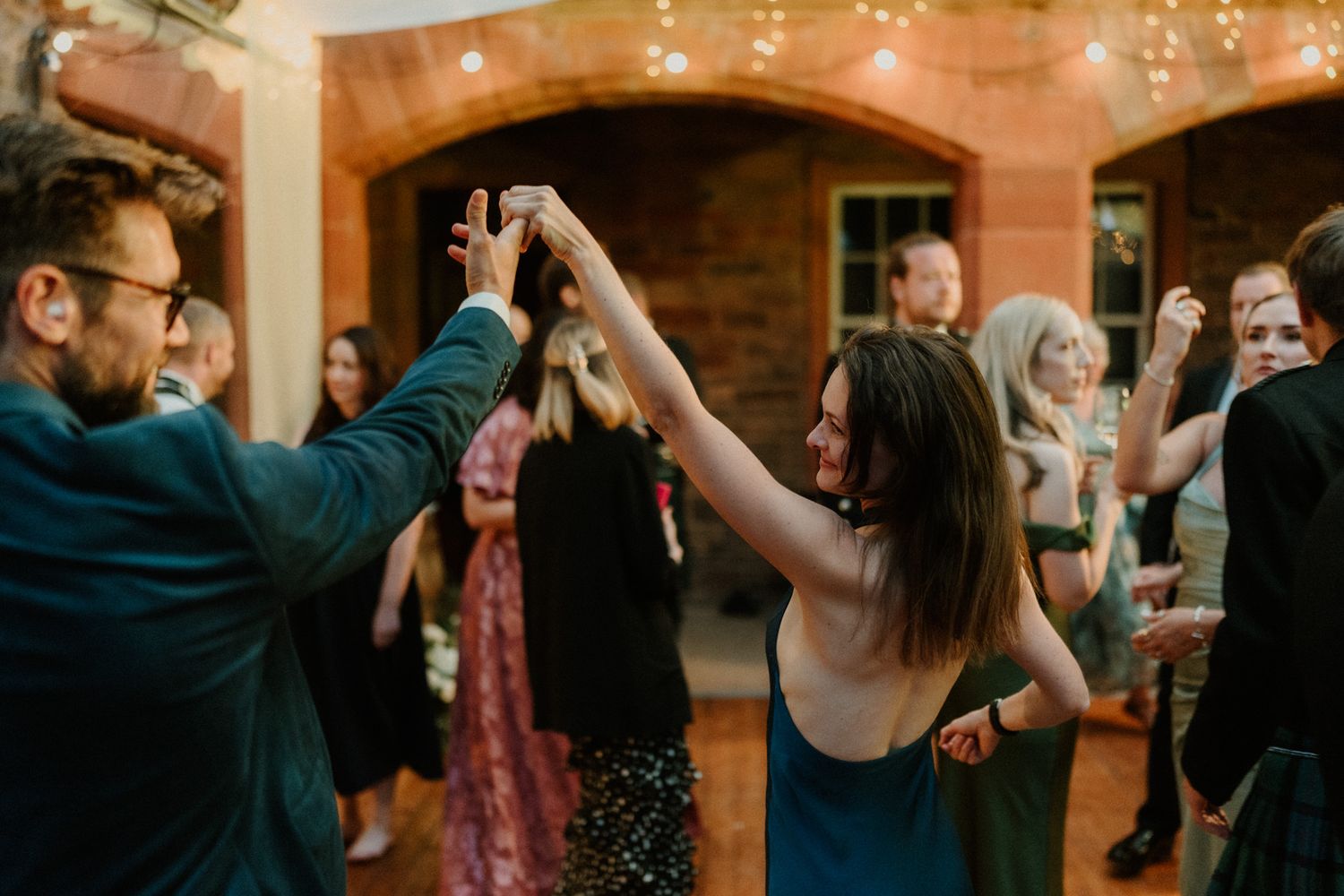 Guests dance and clap at an outdoor evening wedding reception under string lights, with a woman raising her arm in the foreground.