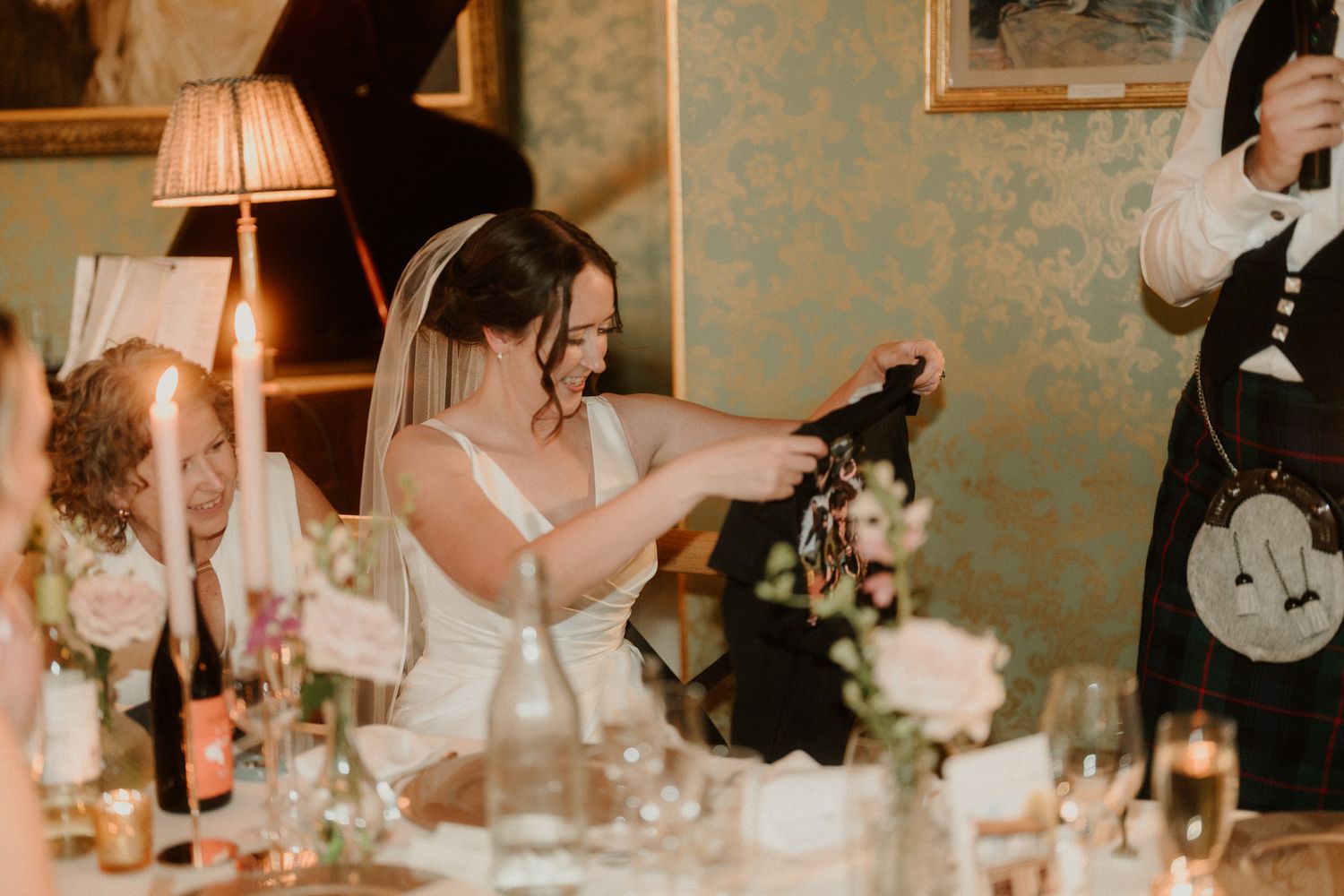 Bride in a white dress and veil seated at a reception table, laughing and holding a dark garment beside candles, flowers and glasses.