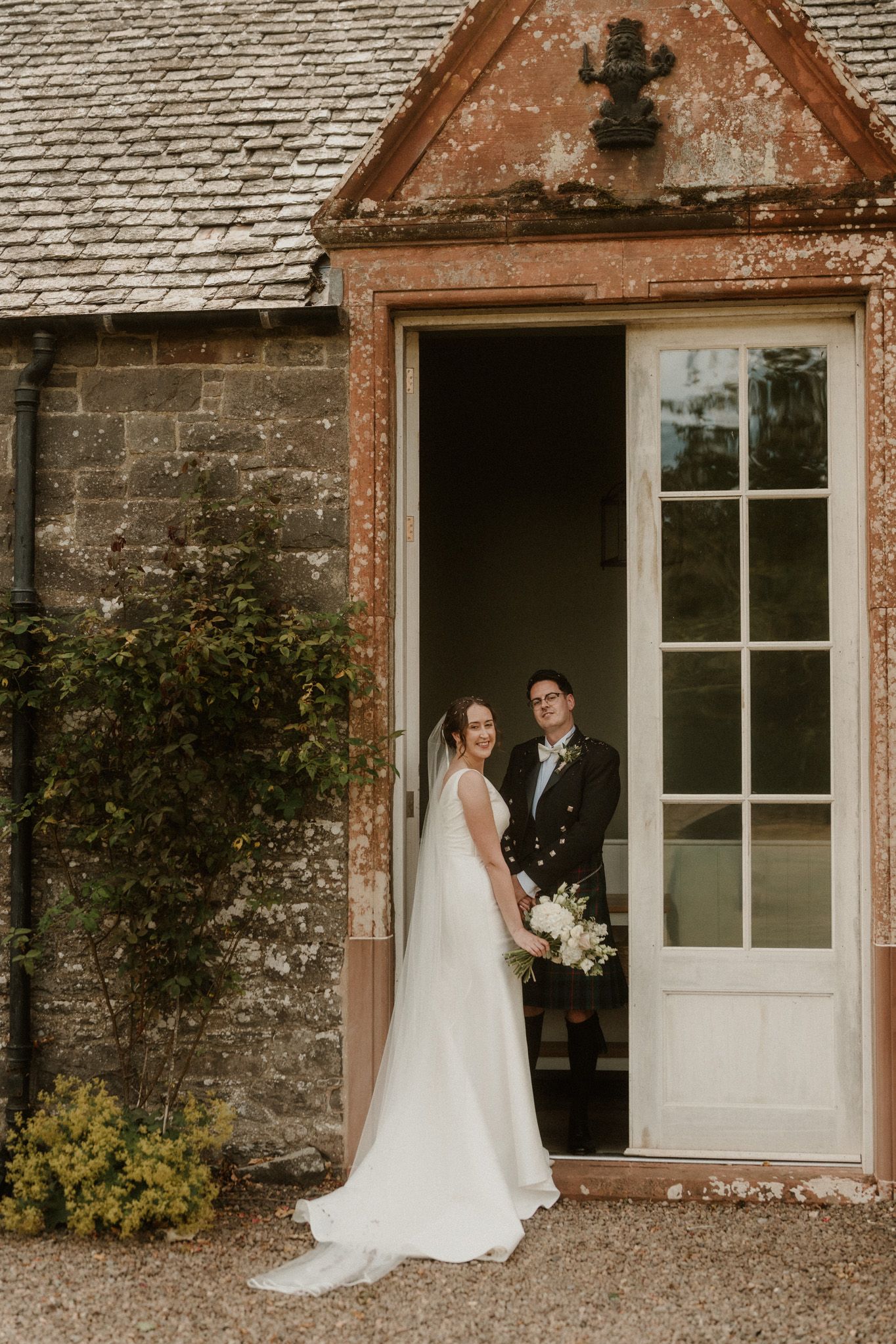 Bride in a flowing white gown holding a bouquet stands beside a groom in a dark jacket in a weathered stone doorway.