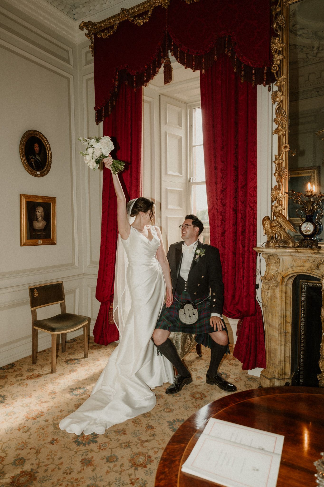 Bride in a white gown raising a bouquet beside a groom in a kilt and black jacket in an elegant room with red velvet curtains.