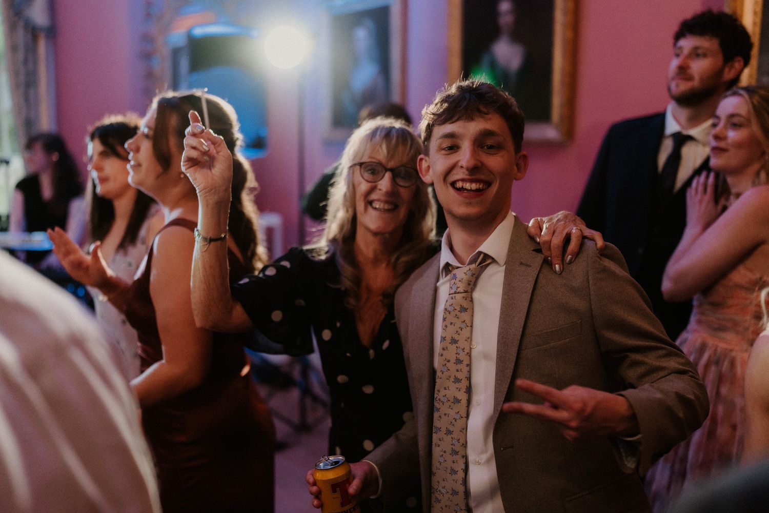 Two guests smile and dance together at a wedding reception in an elegant pink-walled venue.