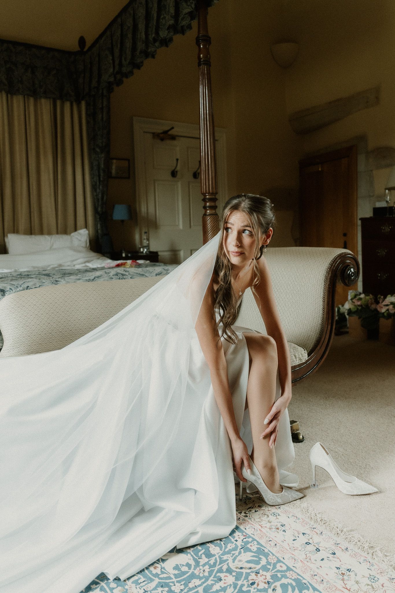 Bride in white wedding dress and veil putting on heels while seated on elegant sofa in luxurious bedroom.