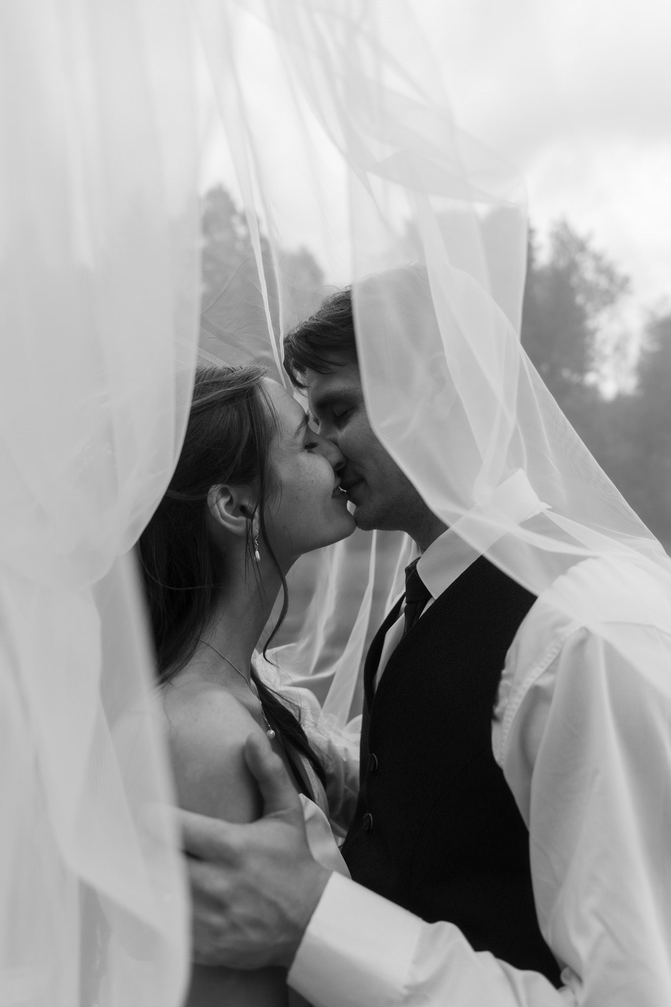 Black and white photo of a bride and groom sharing a romantic kiss beneath a flowing white wedding veil.