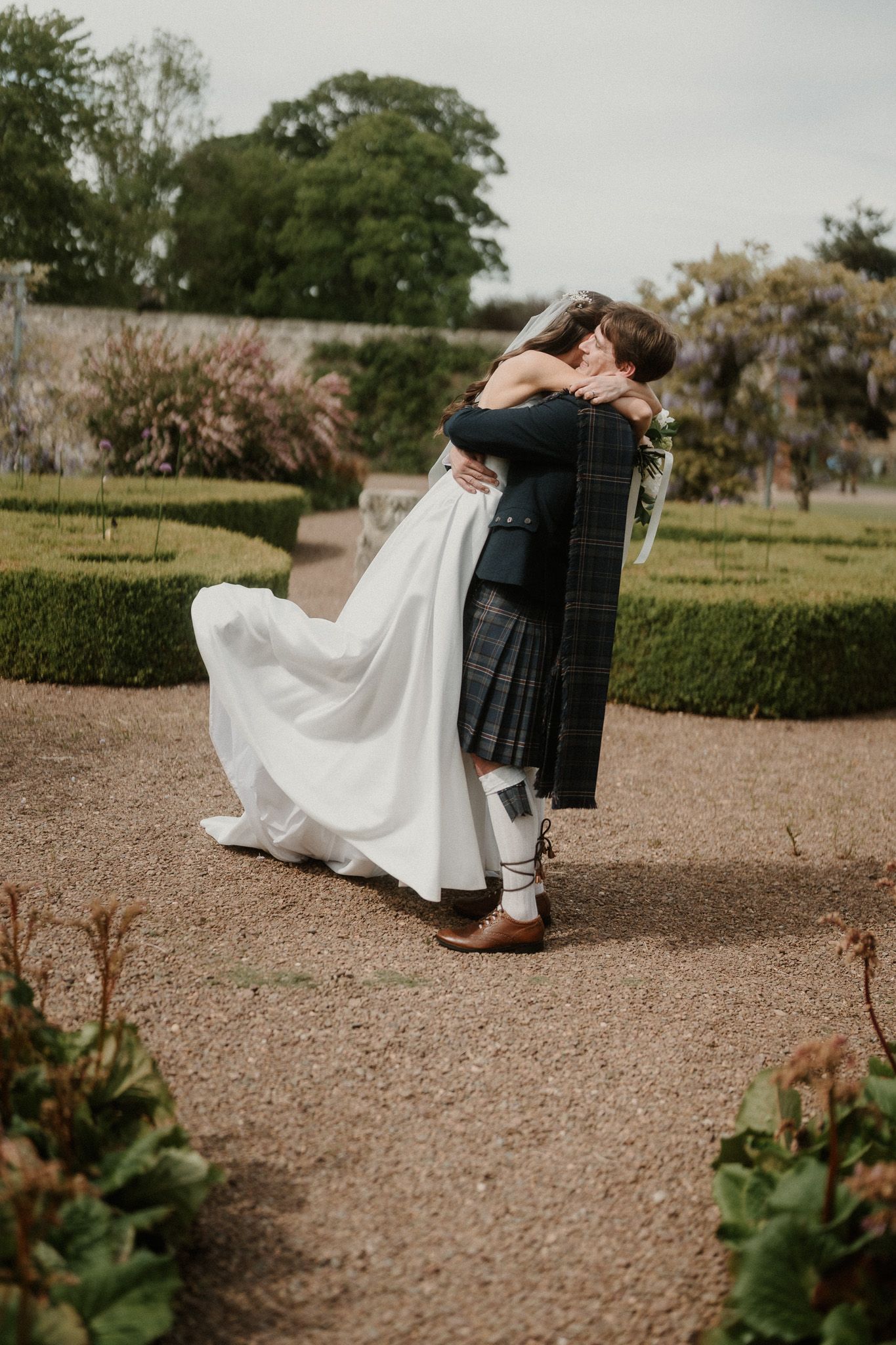 Bride and groom embracing in a formal garden, groom wearing a traditional Scottish kilt and tartan sash.