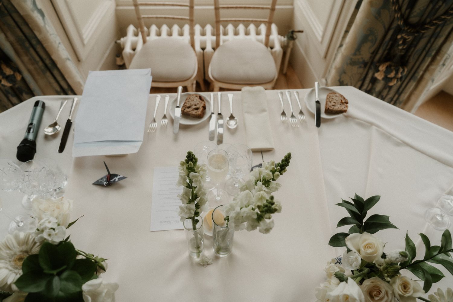 Elegant wedding table setup with white floral arrangements, candles, silverware, and place settings in a bright venue.