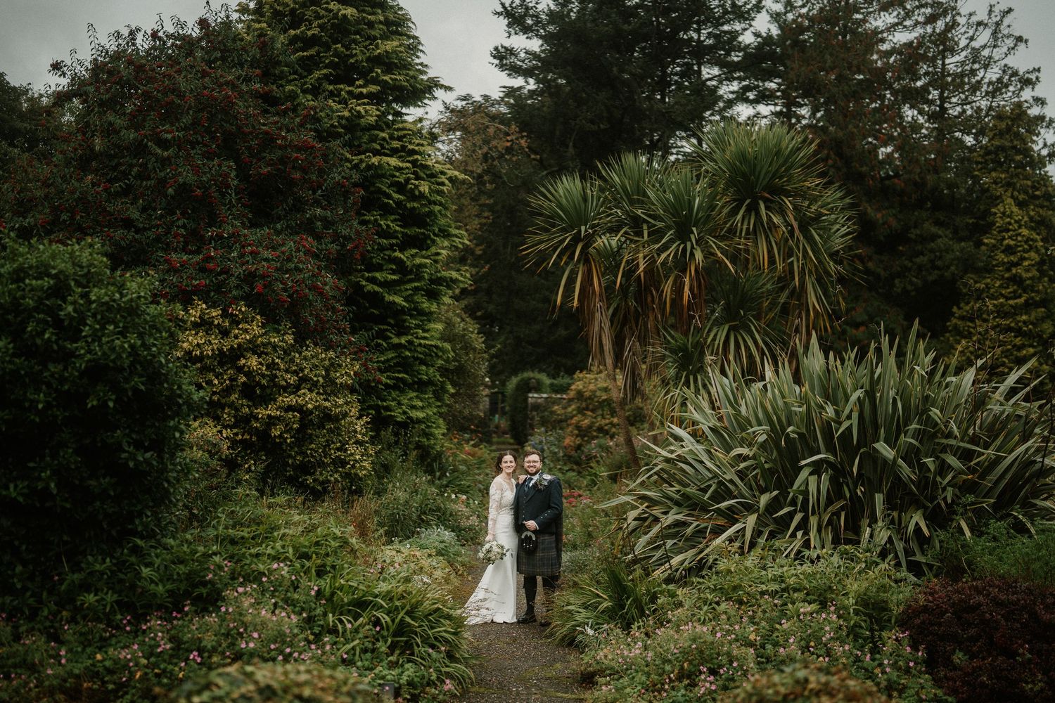 Bride and groom stand together on a garden path surrounded by lush greenery and tropical plants on a cloudy day.