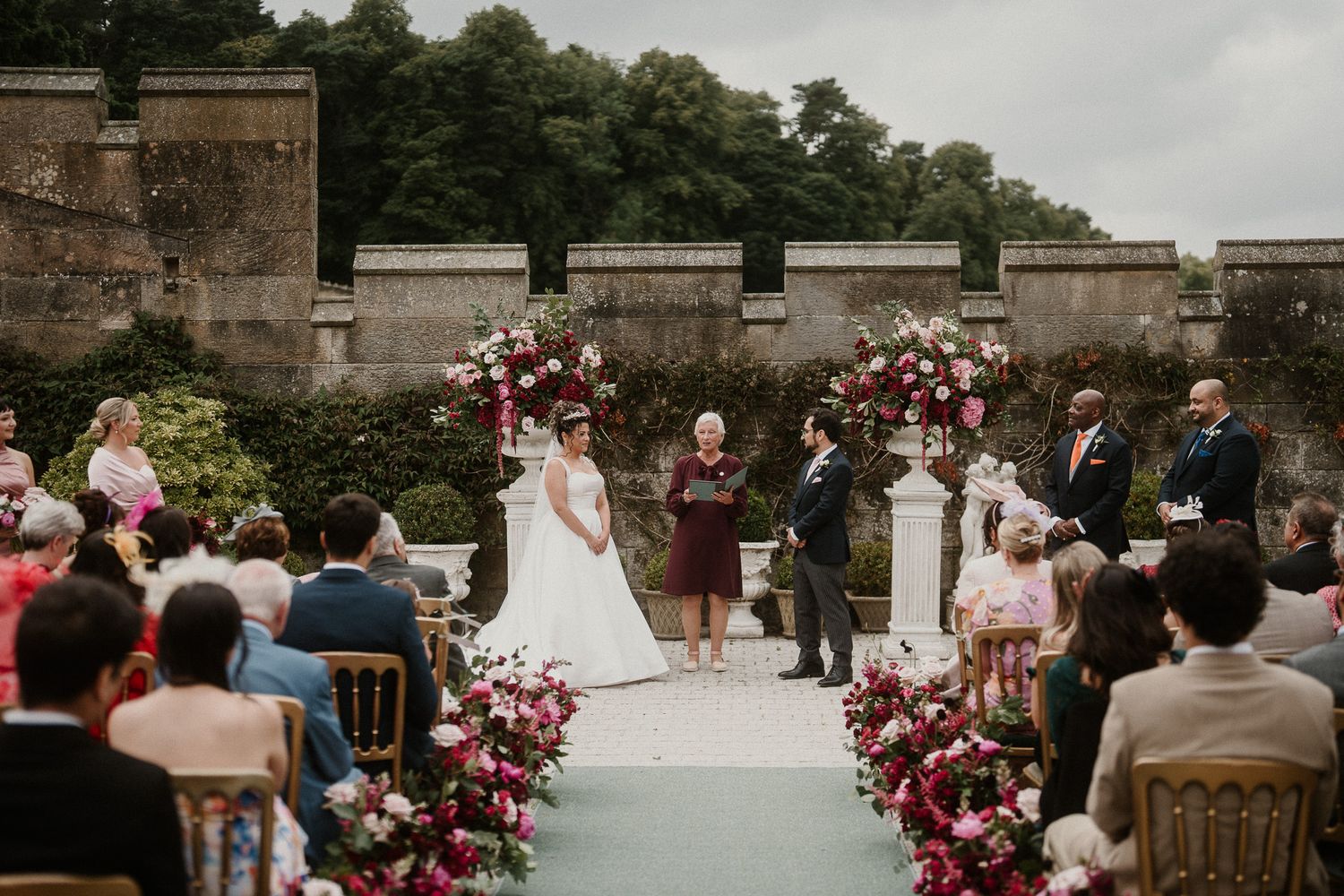 Castle courtyard wedding ceremony with pink floral arrangements and wooden chairs.