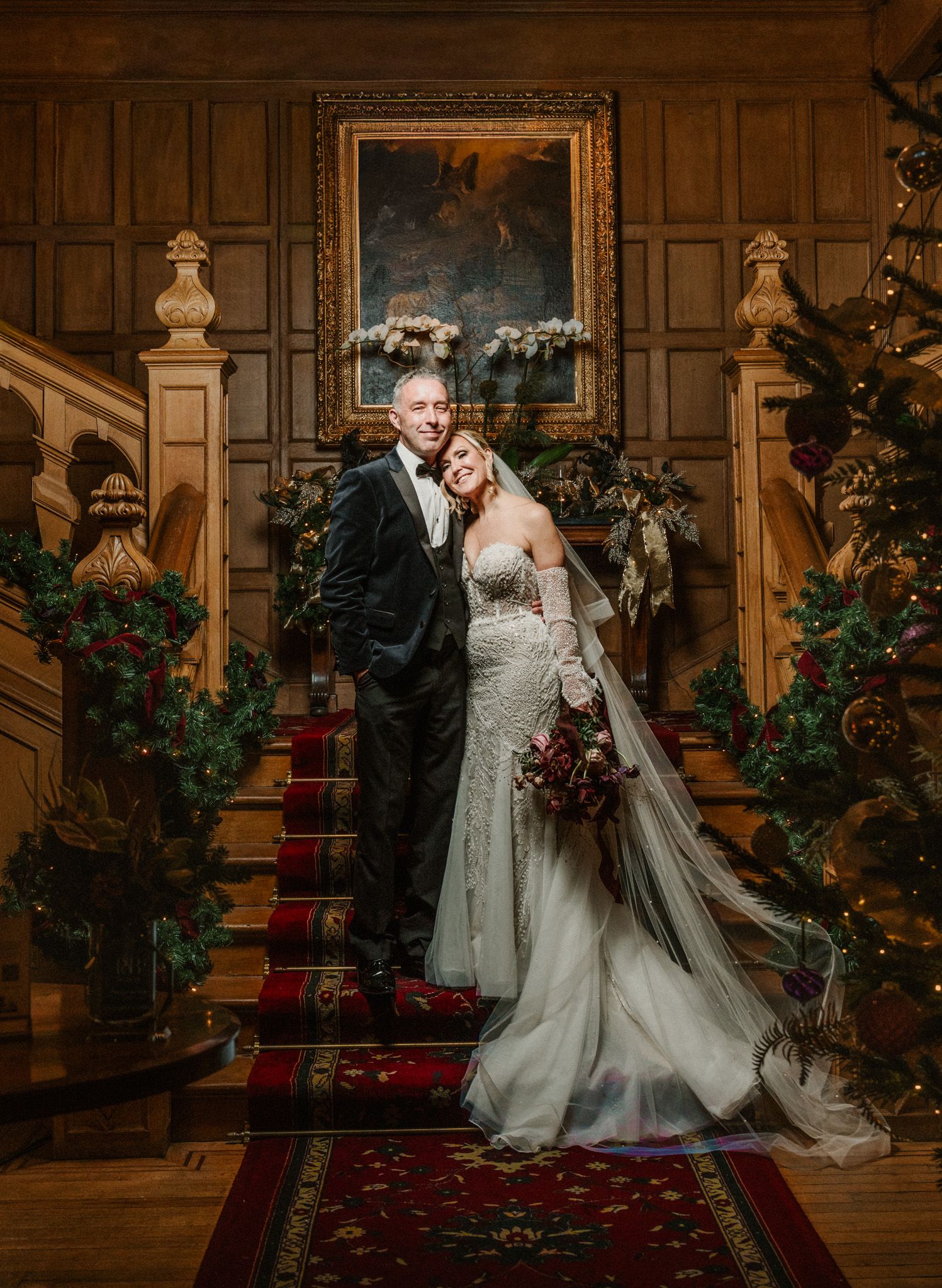 Elegant wedding portrait on grand wooden staircase with red carpet and ornate decorations.