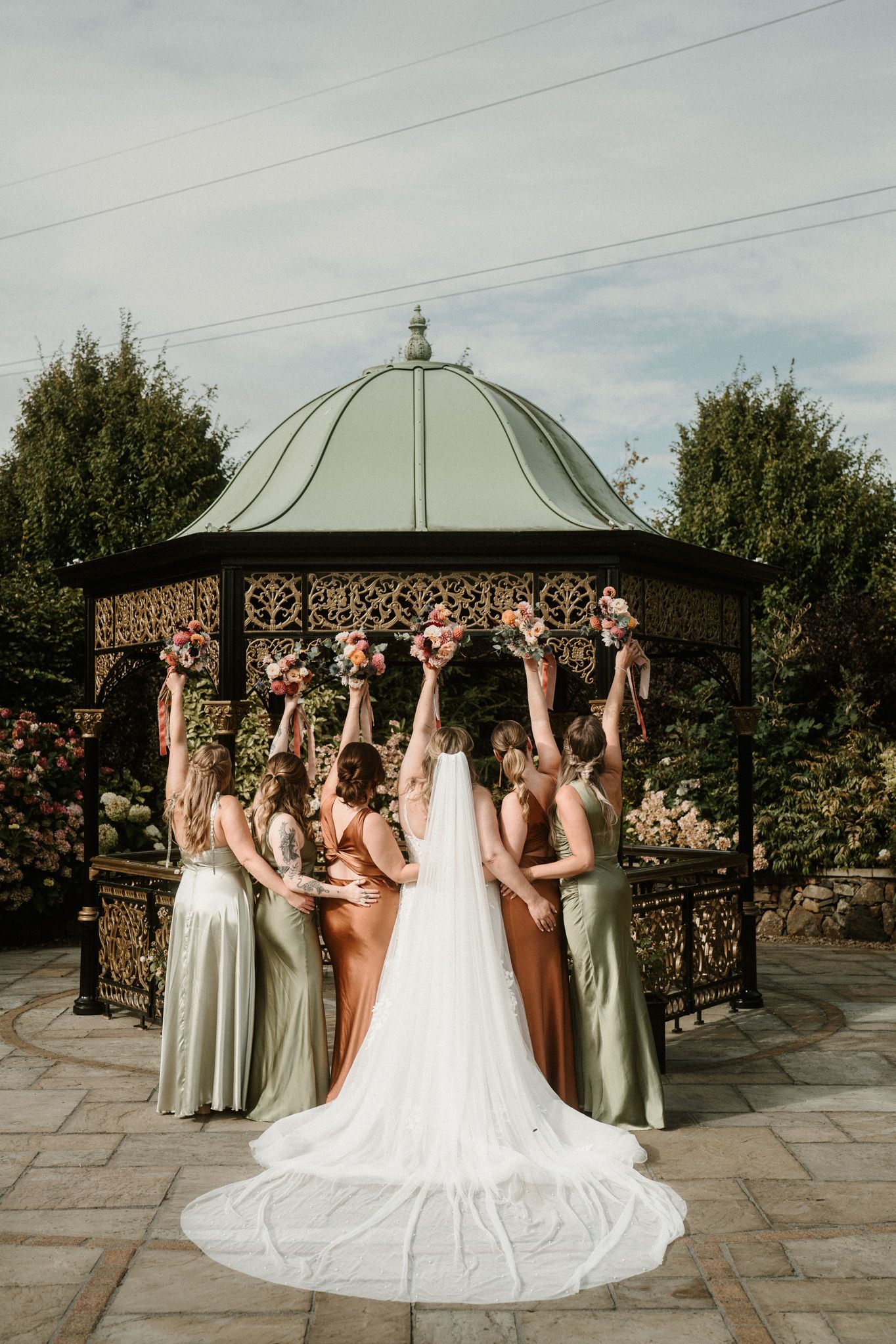 Wedding party celebrating with raised arms under an ornate metal gazebo.