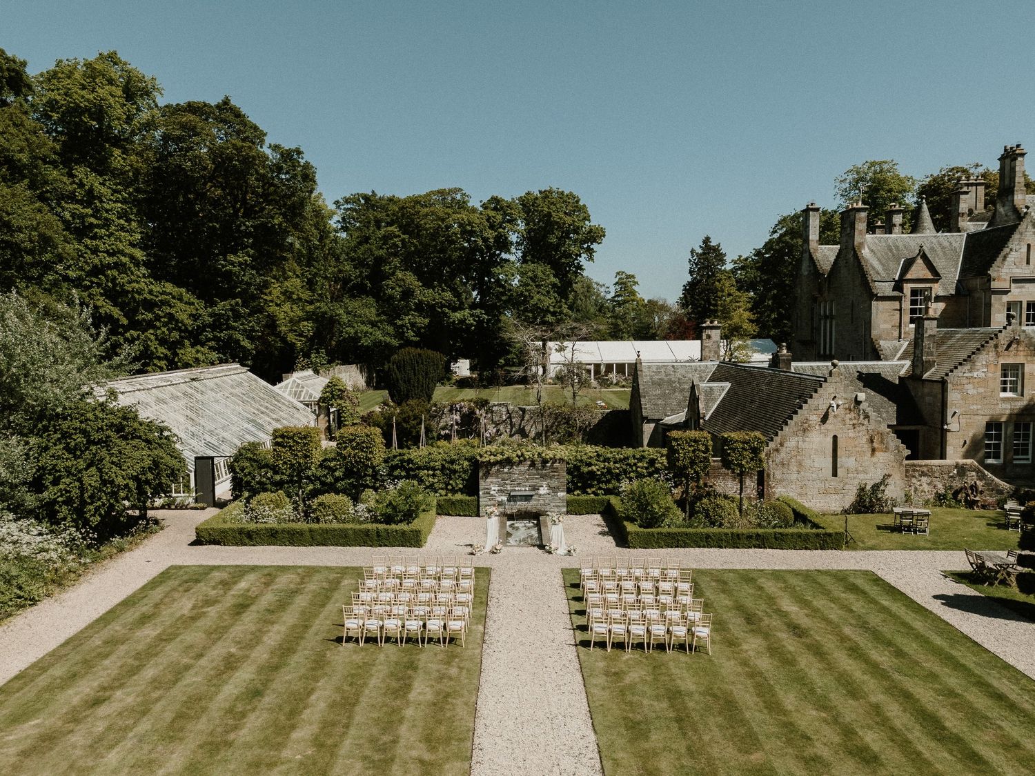 White chairs arranged in rows for an outdoor wedding ceremony with stone buildings and trees in the background.