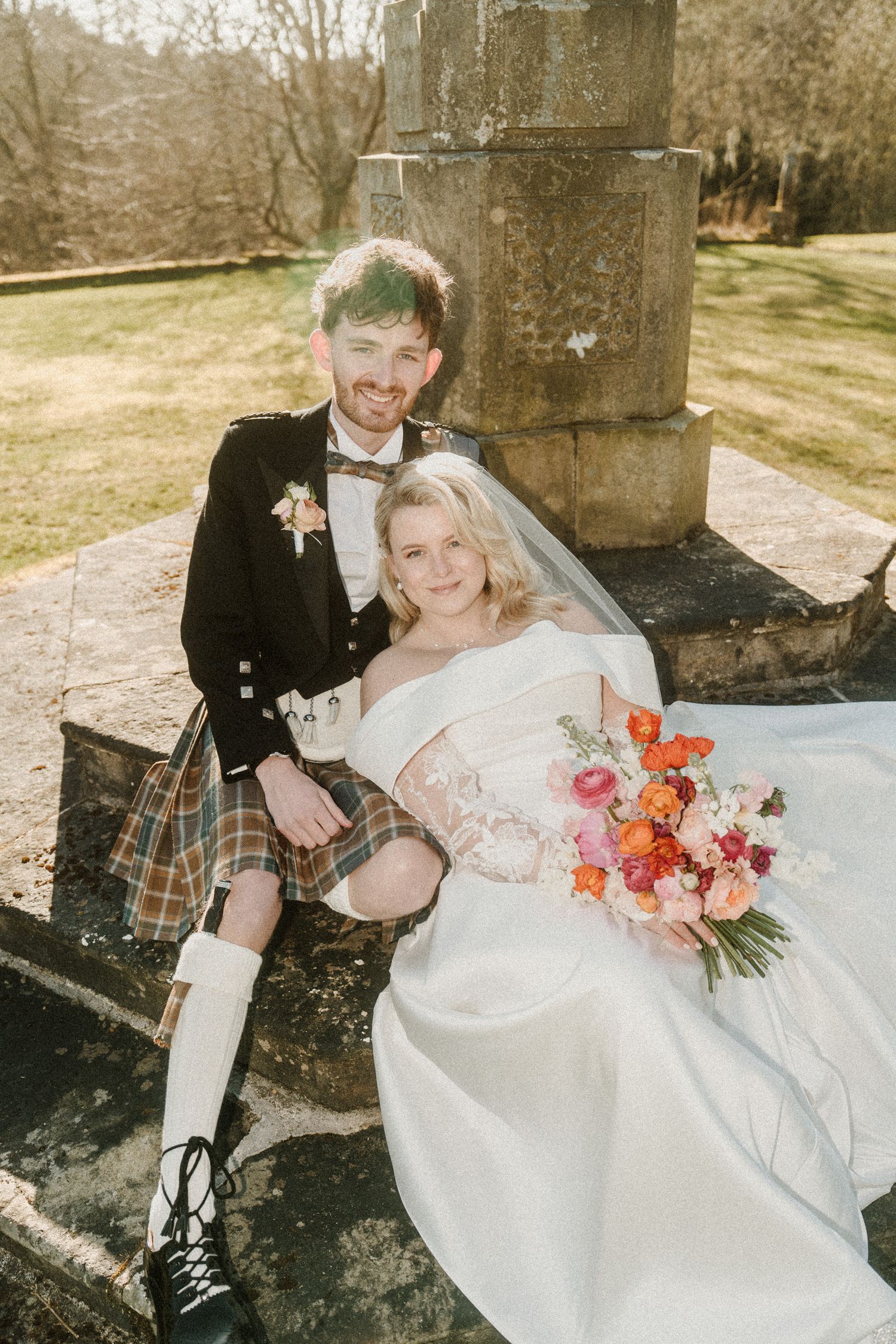 A Scottish wedding couple poses on stone steps, with the groom in a traditional kilt and bride holding coral flowers.