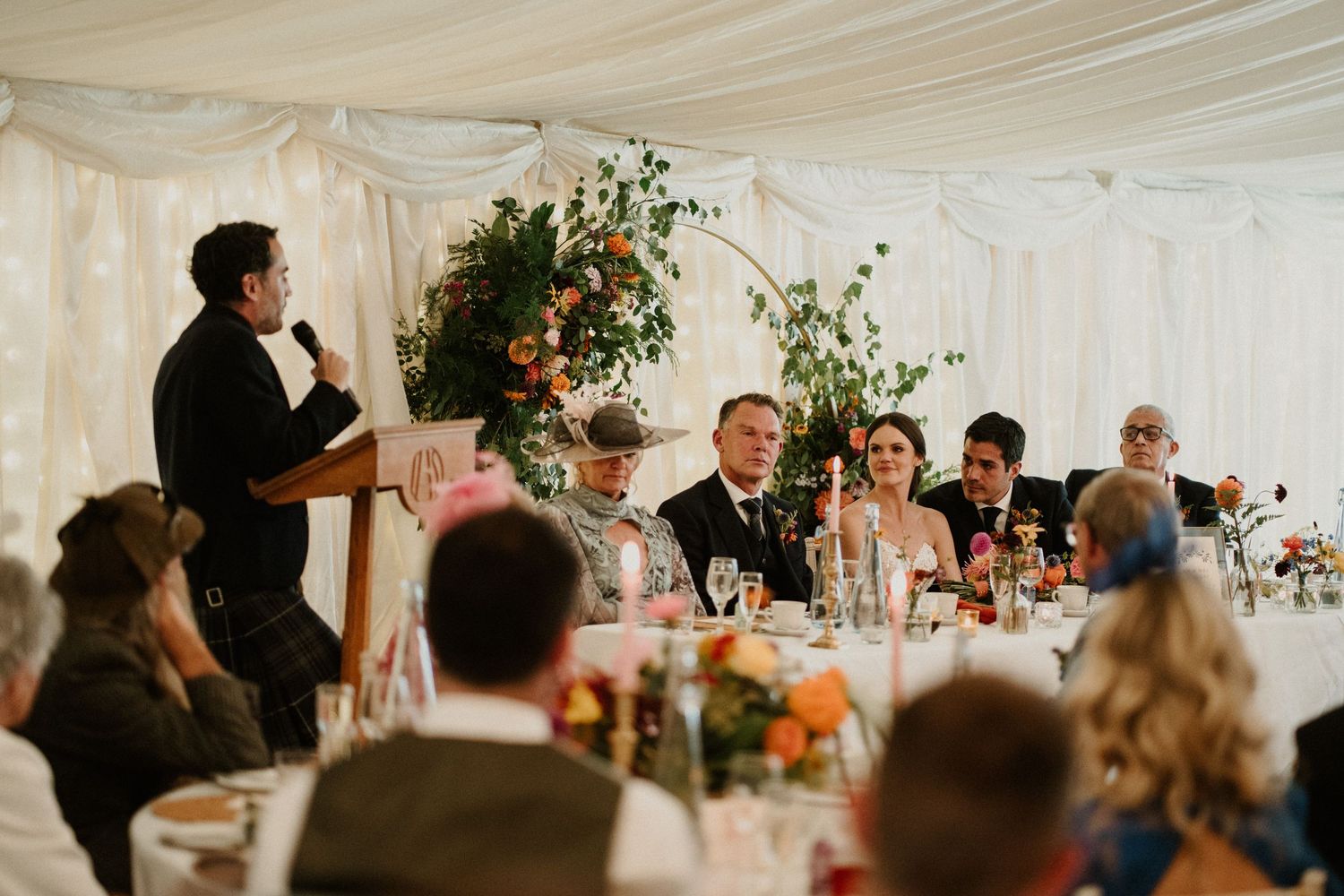 Wedding celebration inside white tent with floral decorations and table settings.