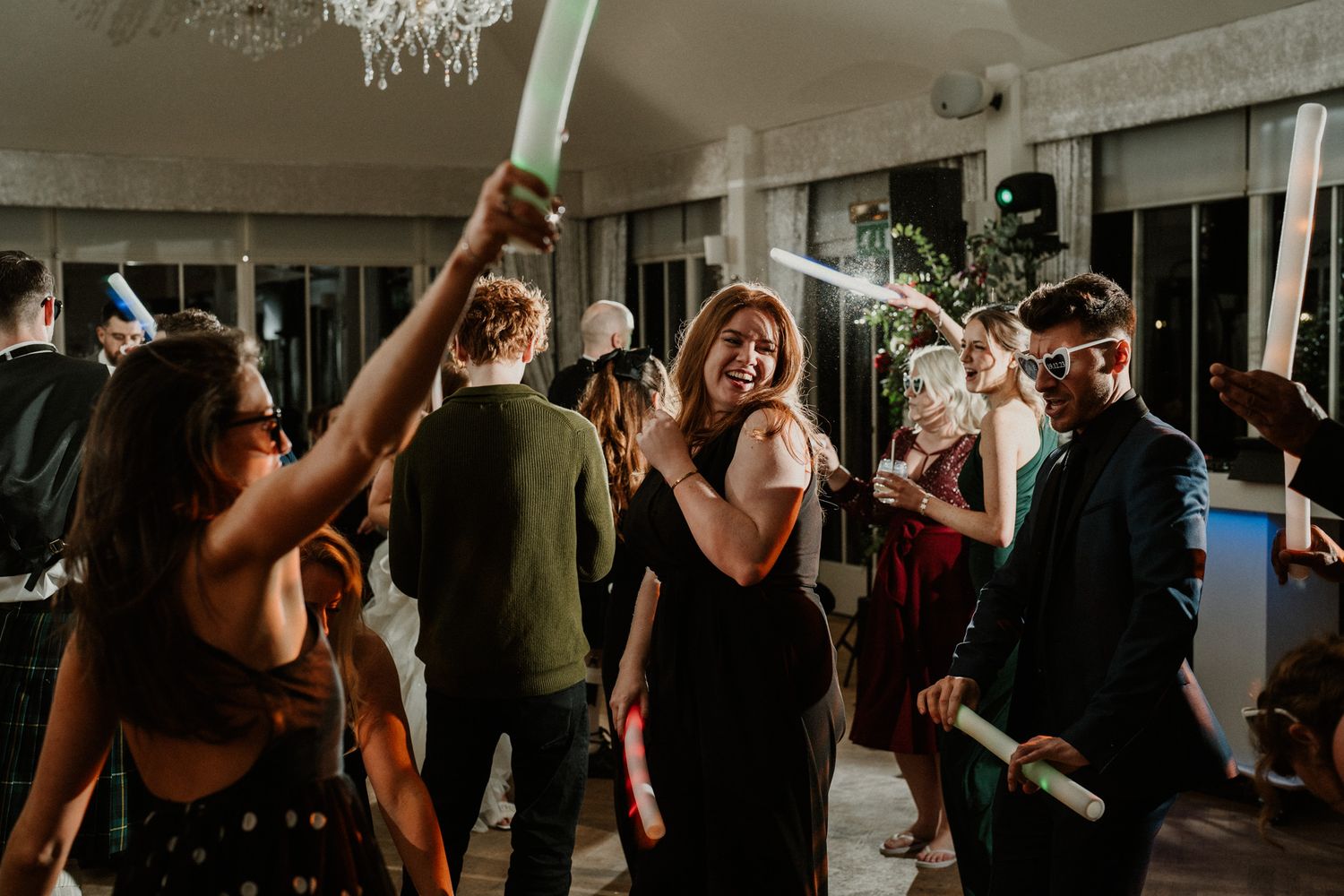 Party guests dancing and celebrating with glow sticks under a chandelier.