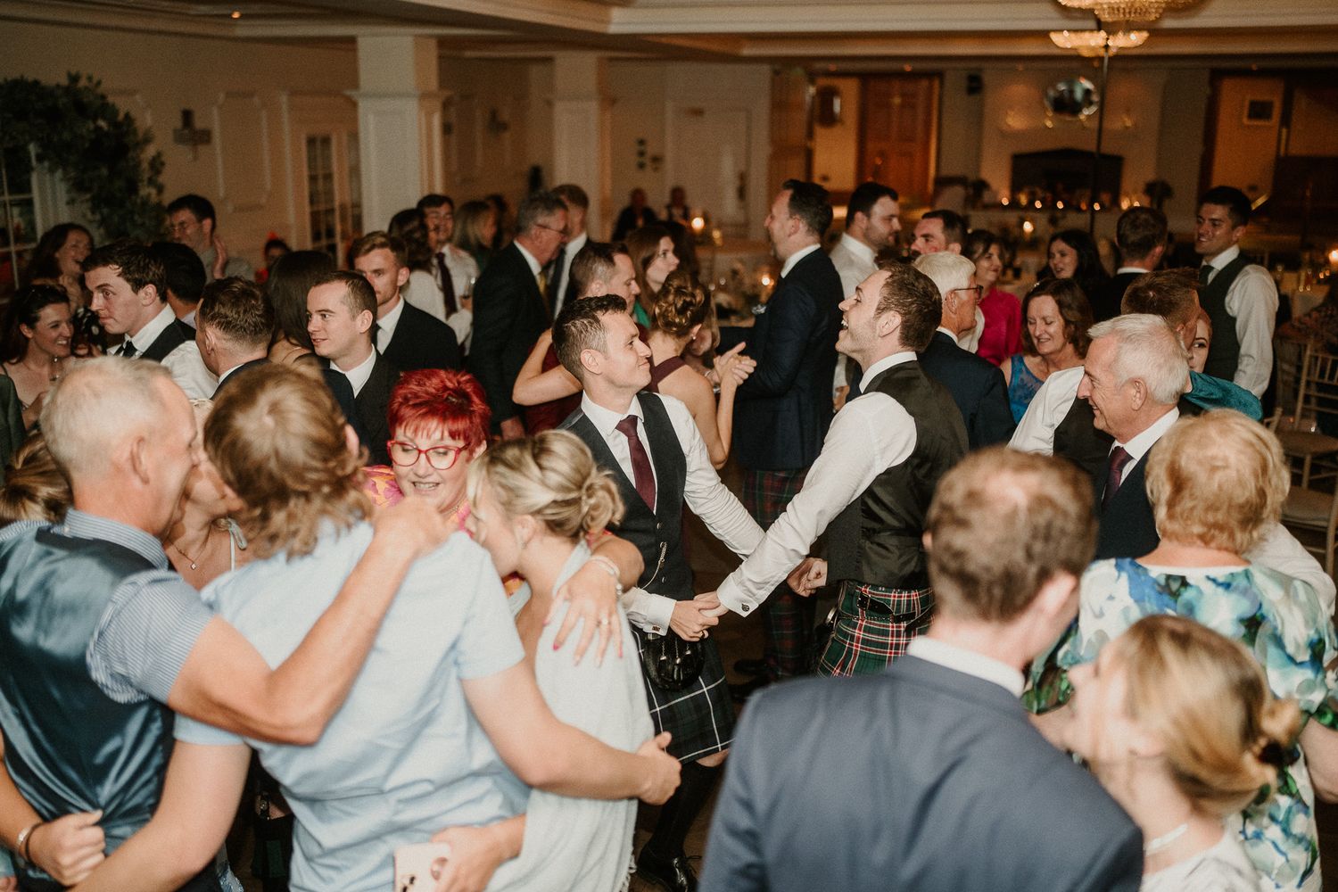Lively wedding reception with couples dancing under ornate chandeliers.