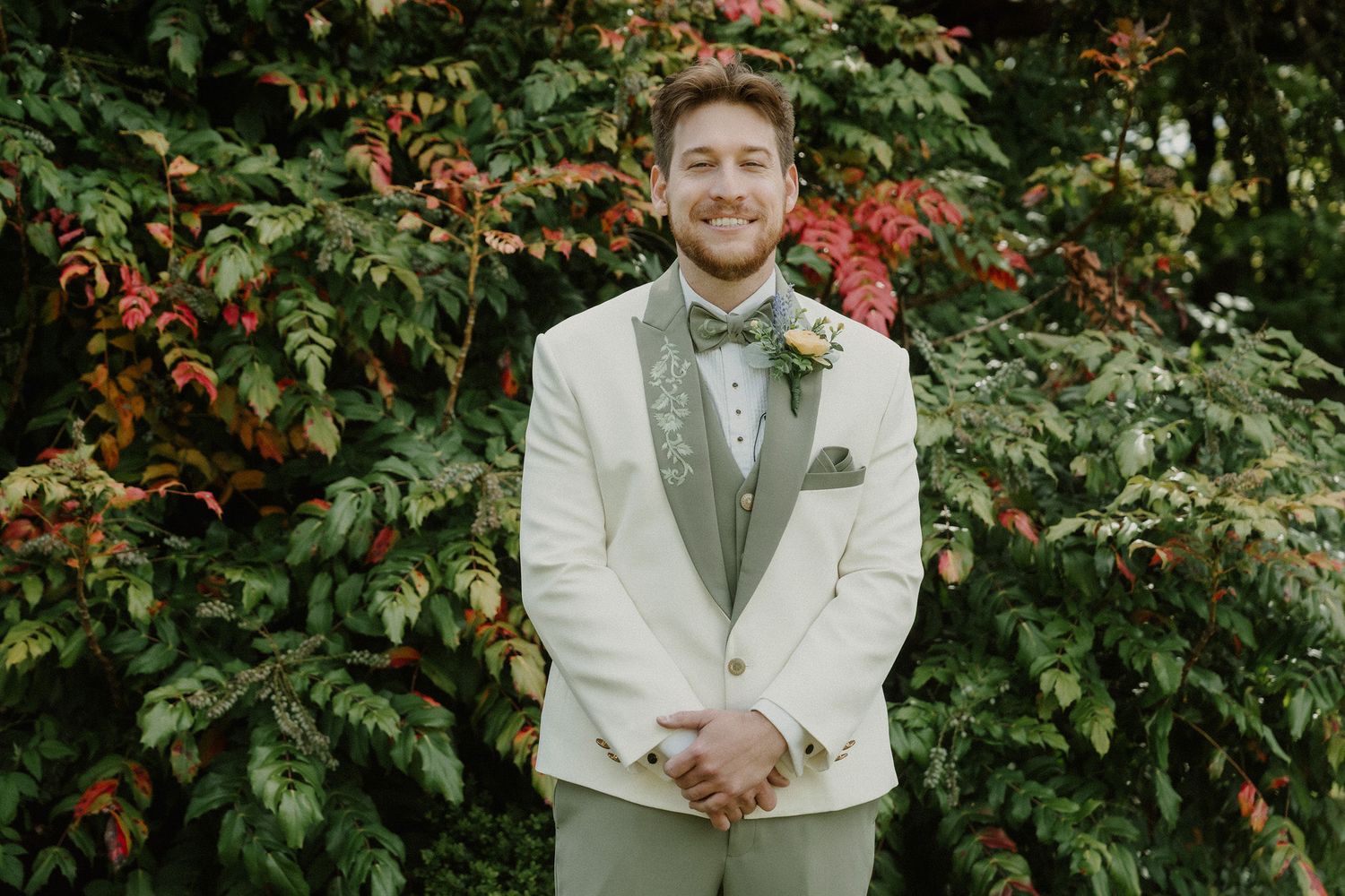 Groom smiling in ivory tuxedo with green lapels and boutonniere, standing before lush green foliage backdrop.