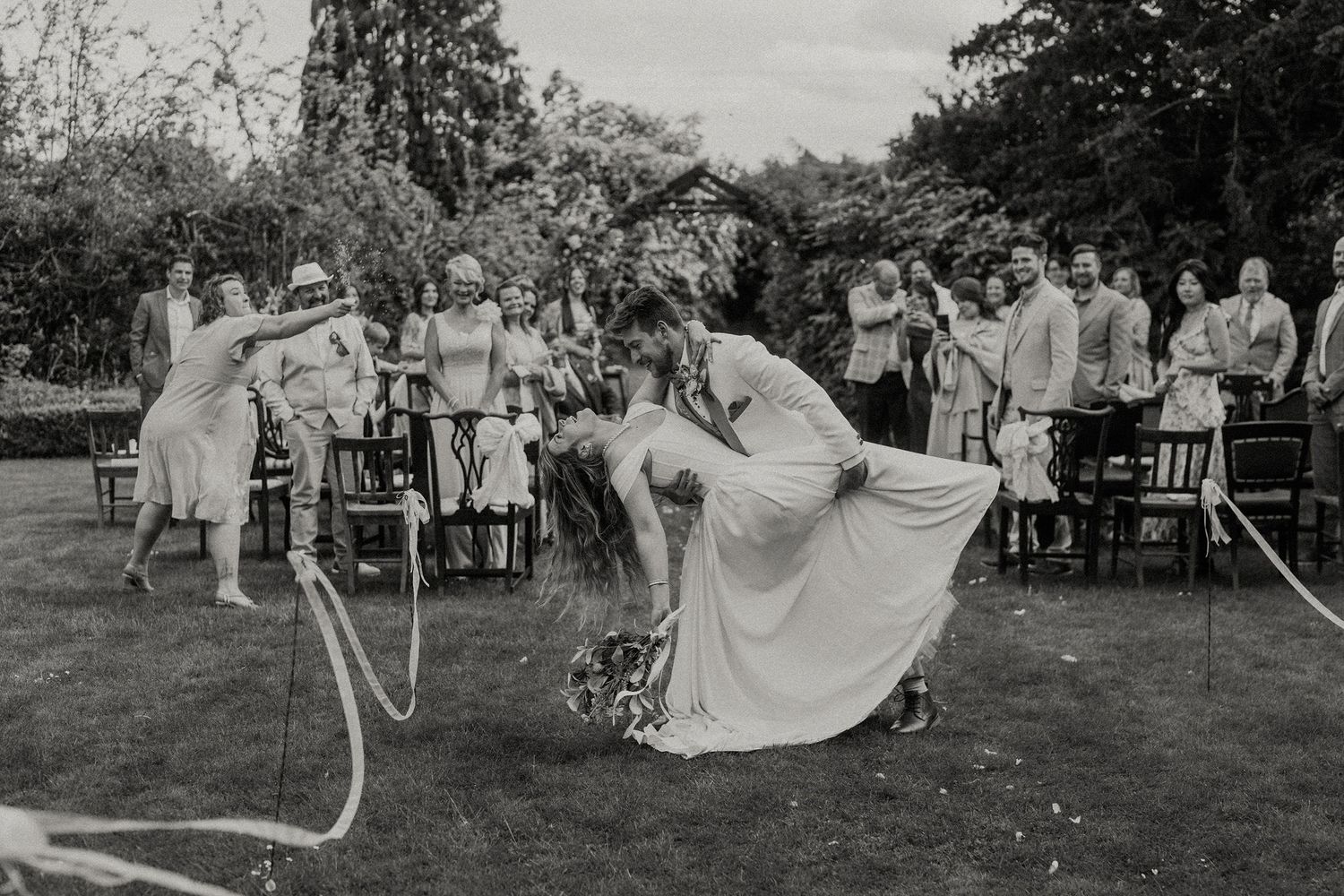 Black and white photo of brides playing limbo with hose at outdoor wedding reception with guests watching.
