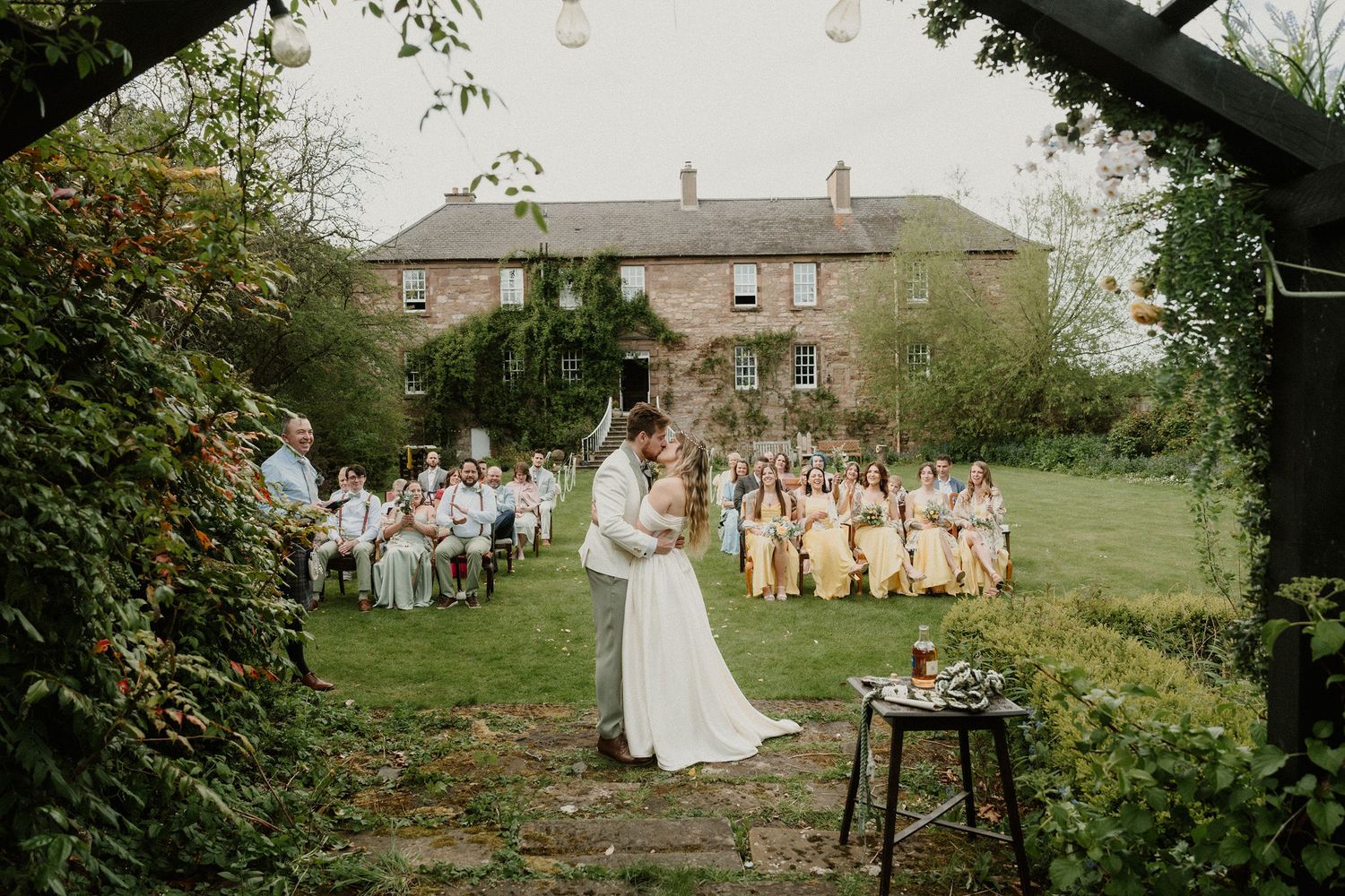 Bride and groom share first dance outdoors at elegant garden wedding with bridesmaids in yellow dresses and historic manor backdrop.