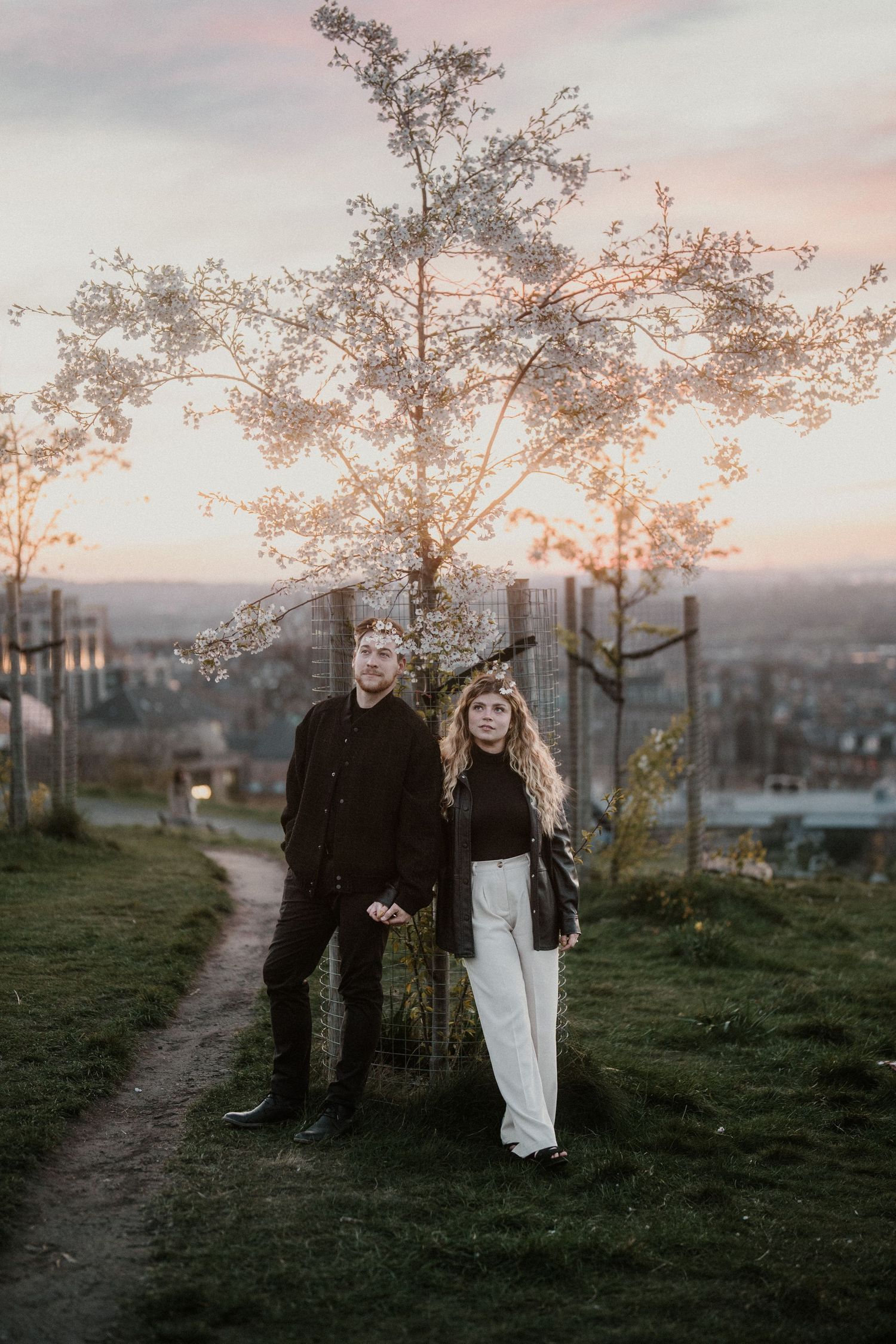 Couple standing before a blossoming tree at sunset with city views behind them.