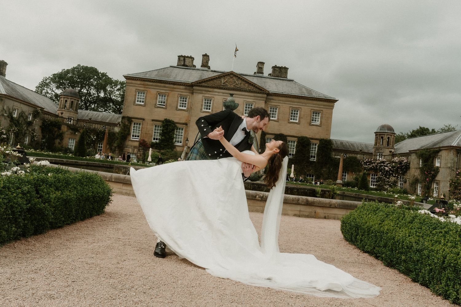 Bride and groom share romantic dip kiss in formal garden outside elegant country house wedding venue.