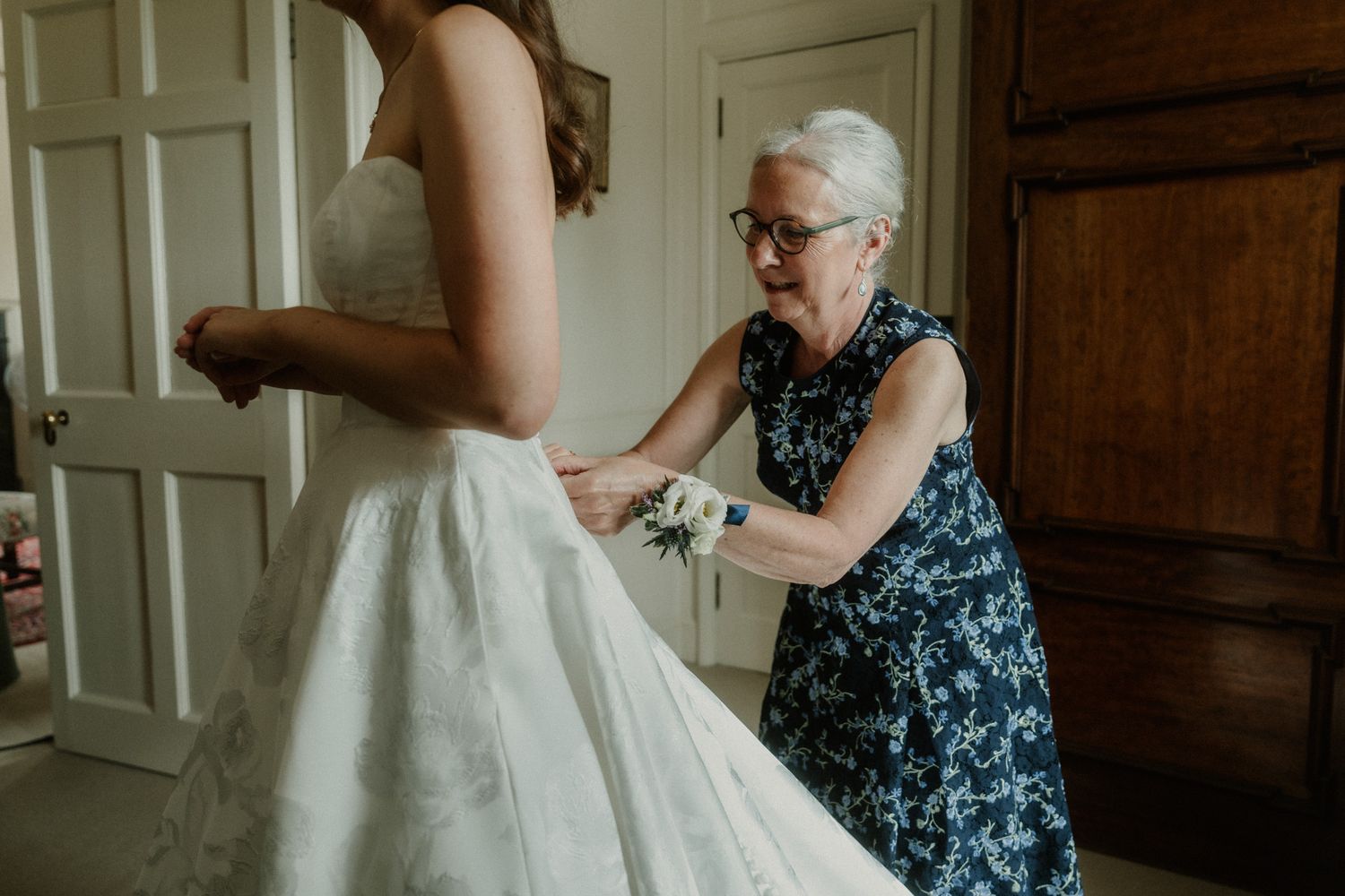 Older woman helping bride button up white wedding dress in elegant room.