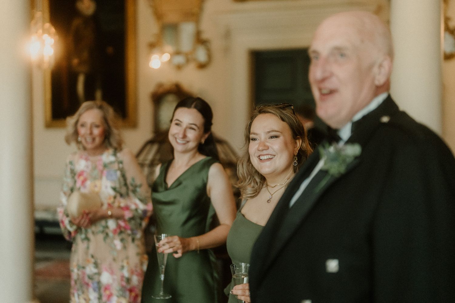 Two bridesmaids in elegant green dresses smile and hold champagne glasses at a wedding reception.
