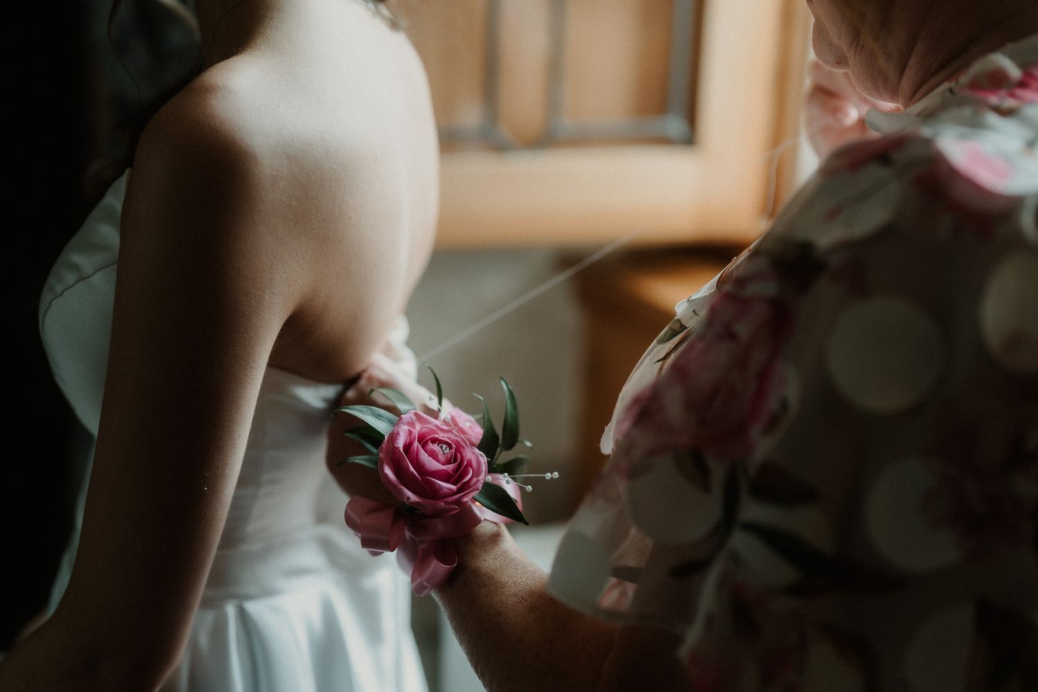 A woman in a white wedding dress receives a pink rose corsage pinned to her gown by another woman in a floral dress.