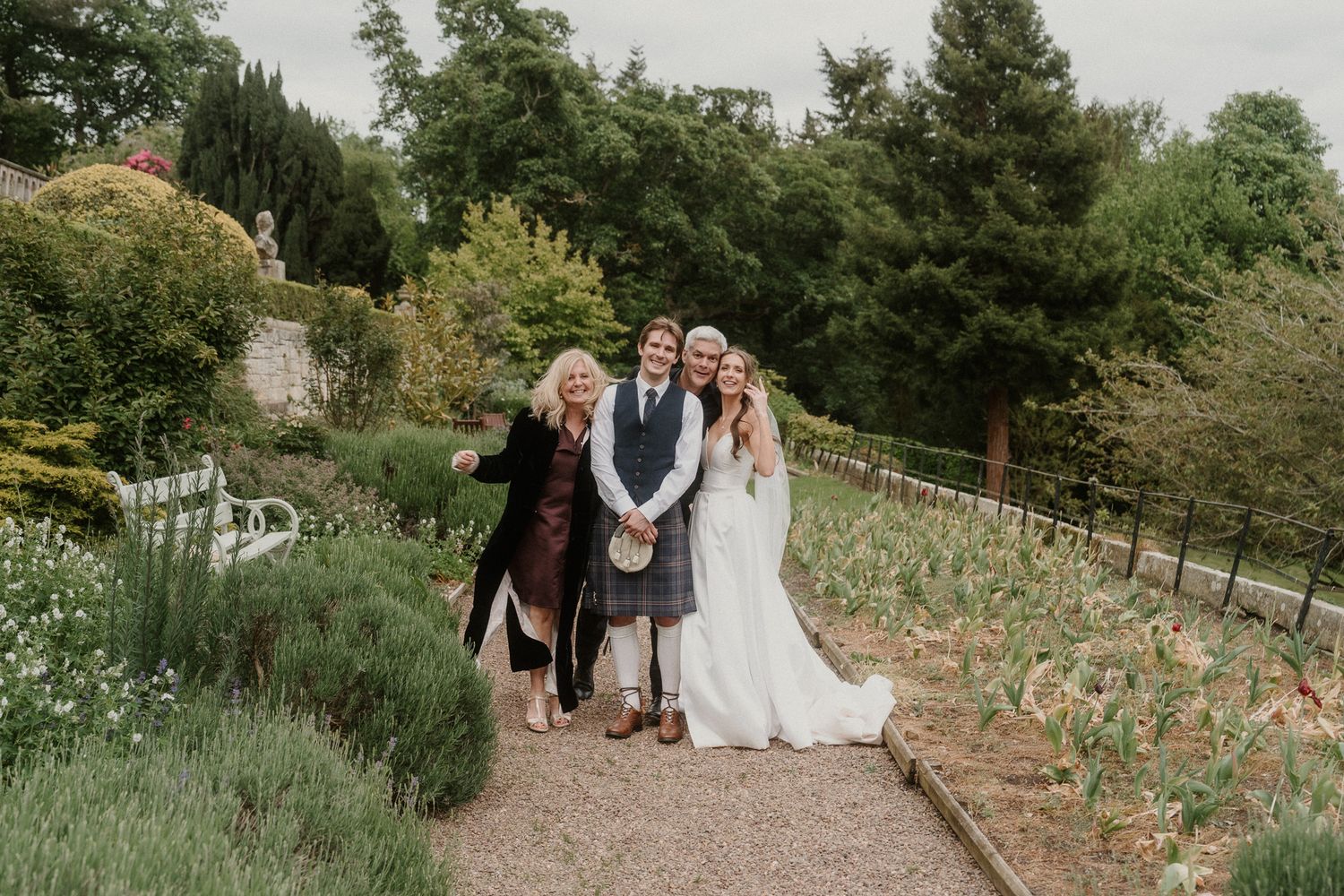 Wedding party posing in a lush garden path, bride in white gown, groom in kilt, with two guests smiling.