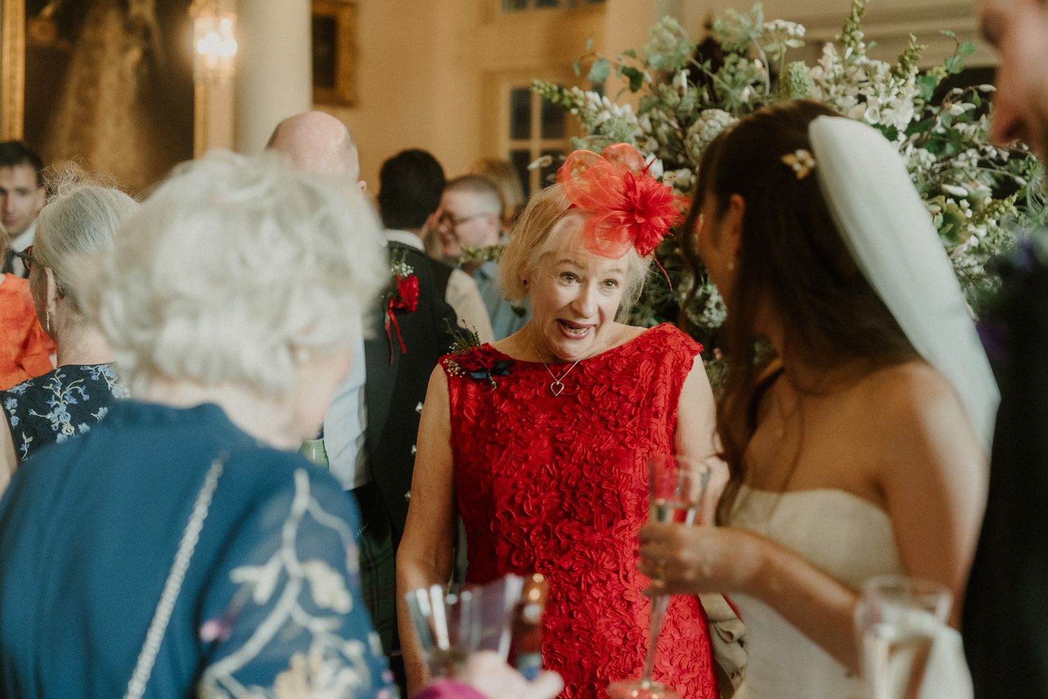 Woman in red lace dress and fascinator smiling at a wedding reception with guests and floral decor.