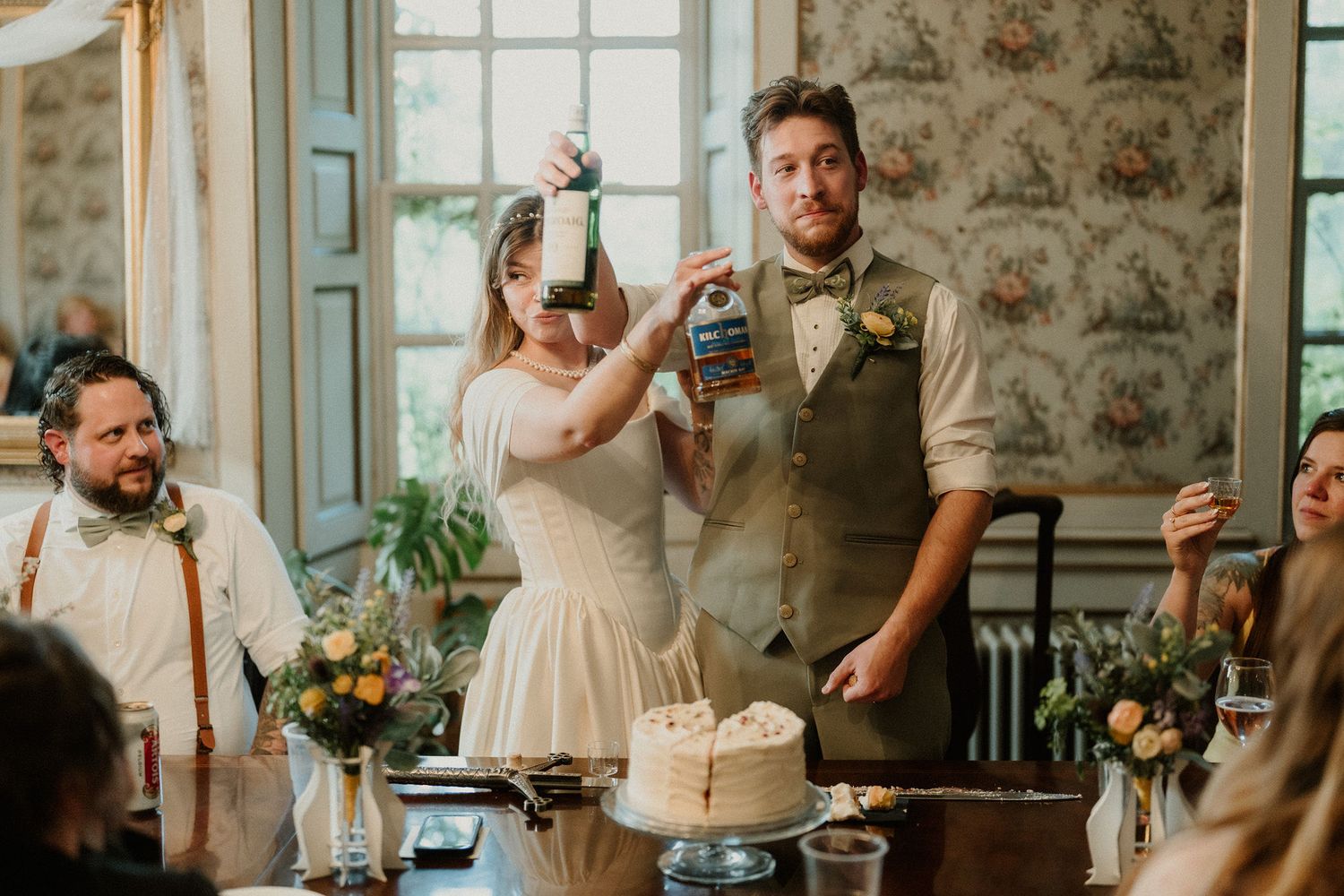Bride and groom cutting wedding cake while holding craft beer bottles at rustic indoor wedding reception.