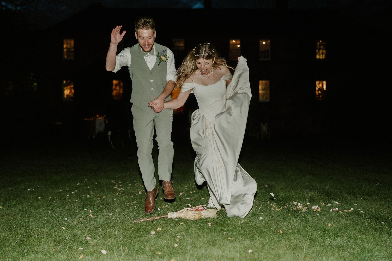 Bride and groom dancing joyfully on grass at night outside an elegant venue, bride in flowing gown.