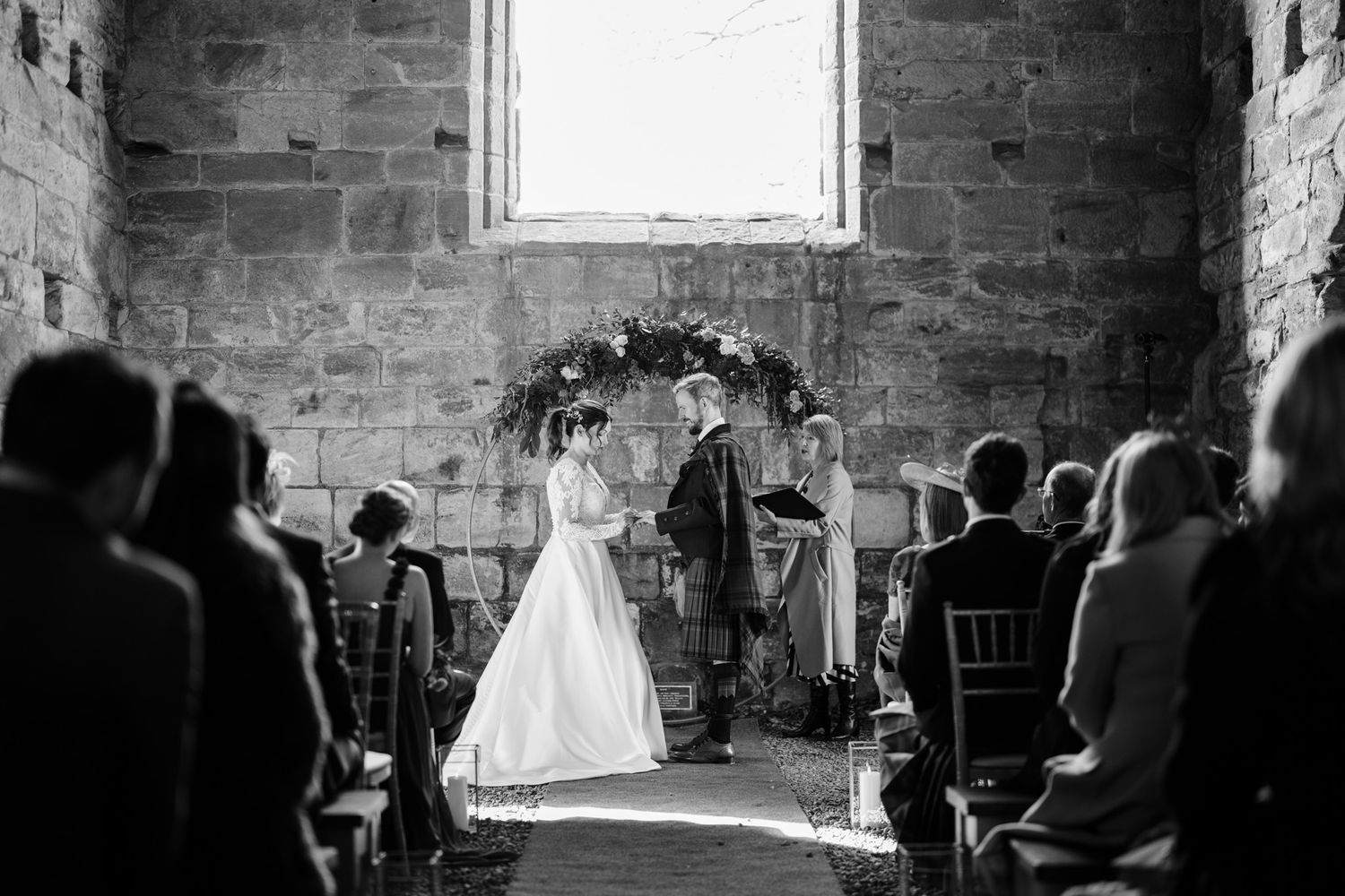 A wedding ceremony in a historic stone chapel with natural light streaming through a window behind the couple.